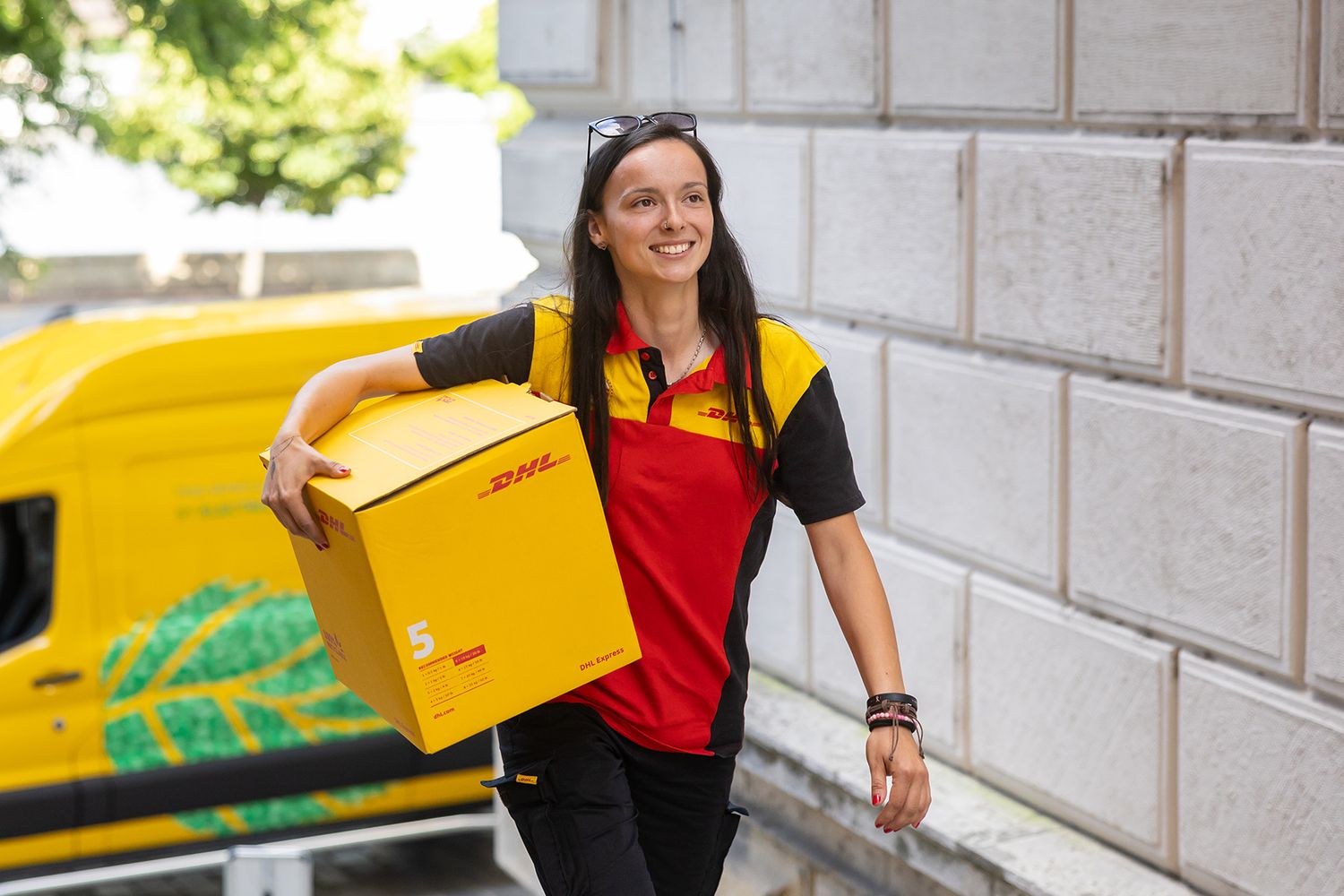 A delivery codestrone carries multiple packages through a garden walkway in a yellow and red uniform.