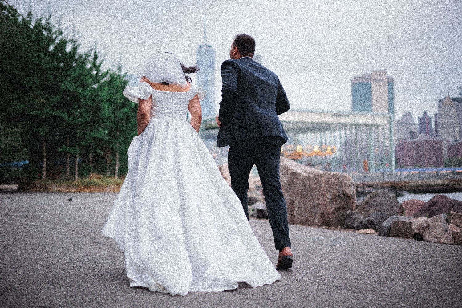Wedding couple walking along waterfront path with city skyline in background.