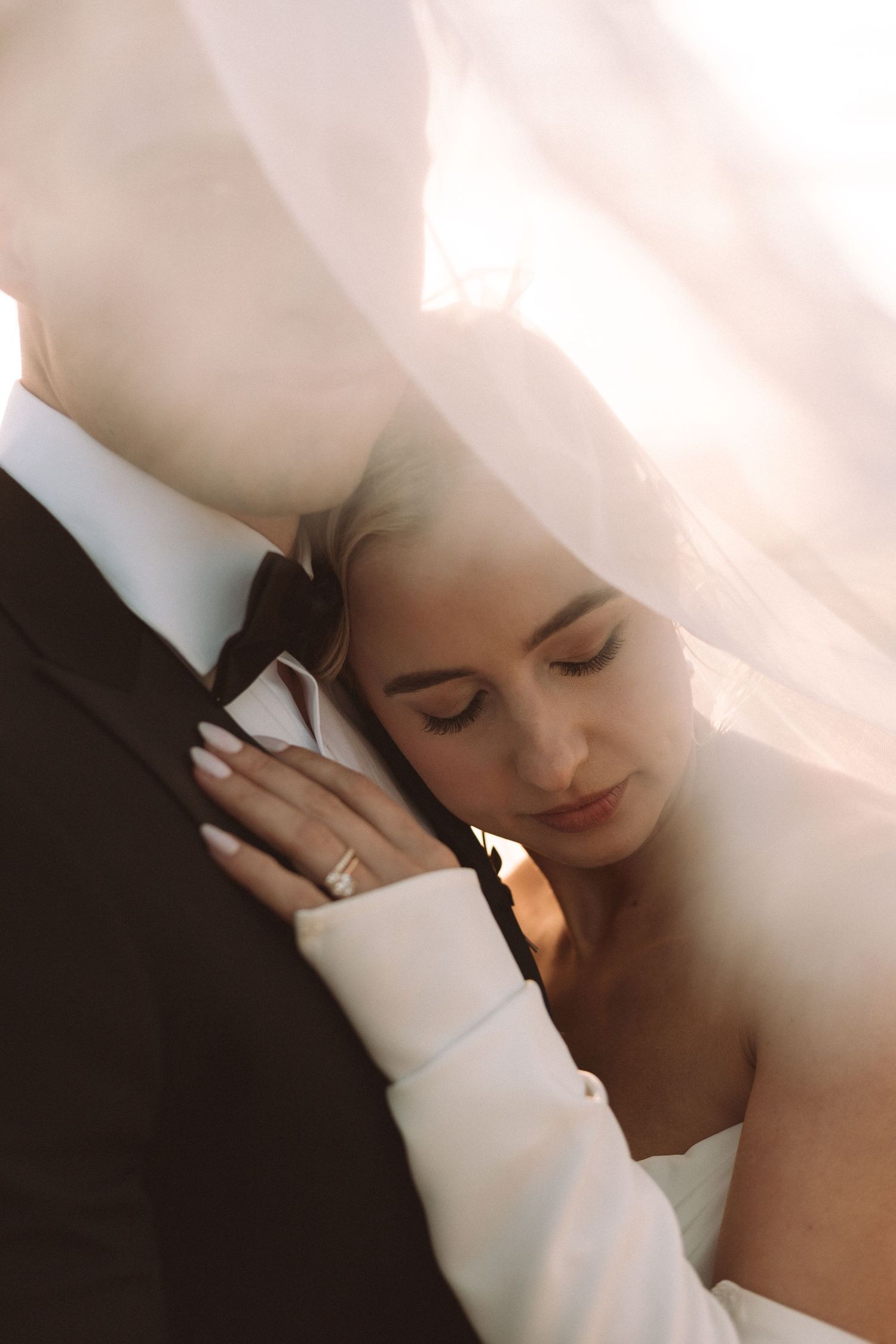 Intimate wedding portrait of couple embracing under soft light with delicate veil and formal attire.