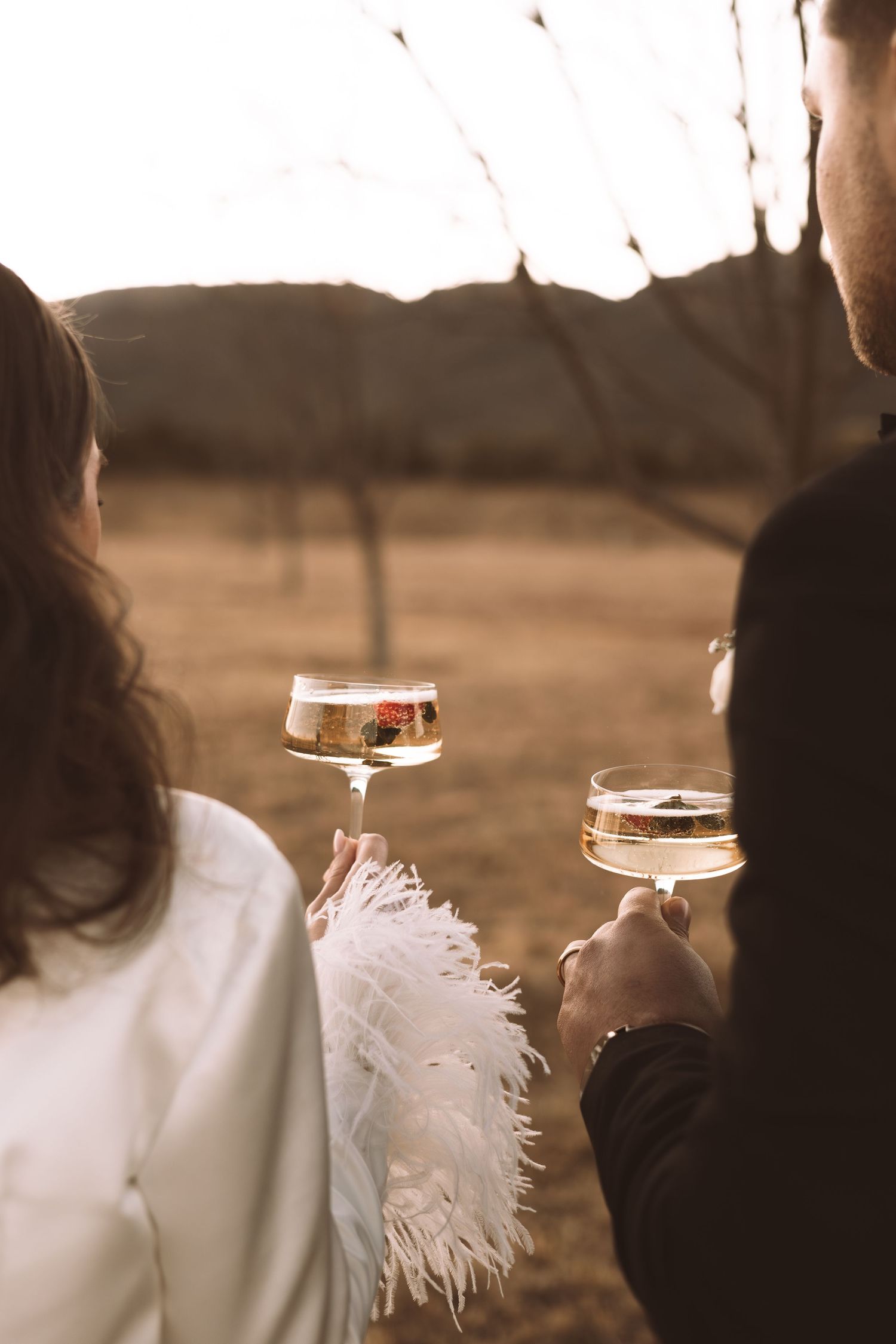 Couple toasting with champagne glasses at sunset wearing wedding attire.