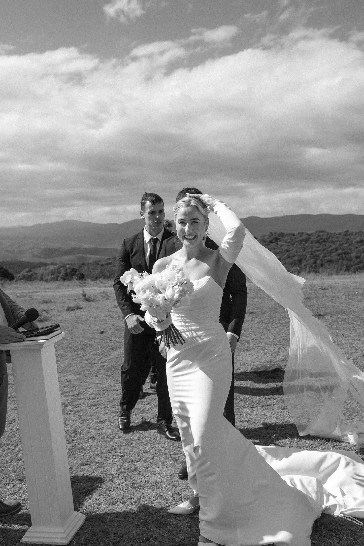 A wedding couple poses outdoors in a field with dramatic clouds and mountains in the background.