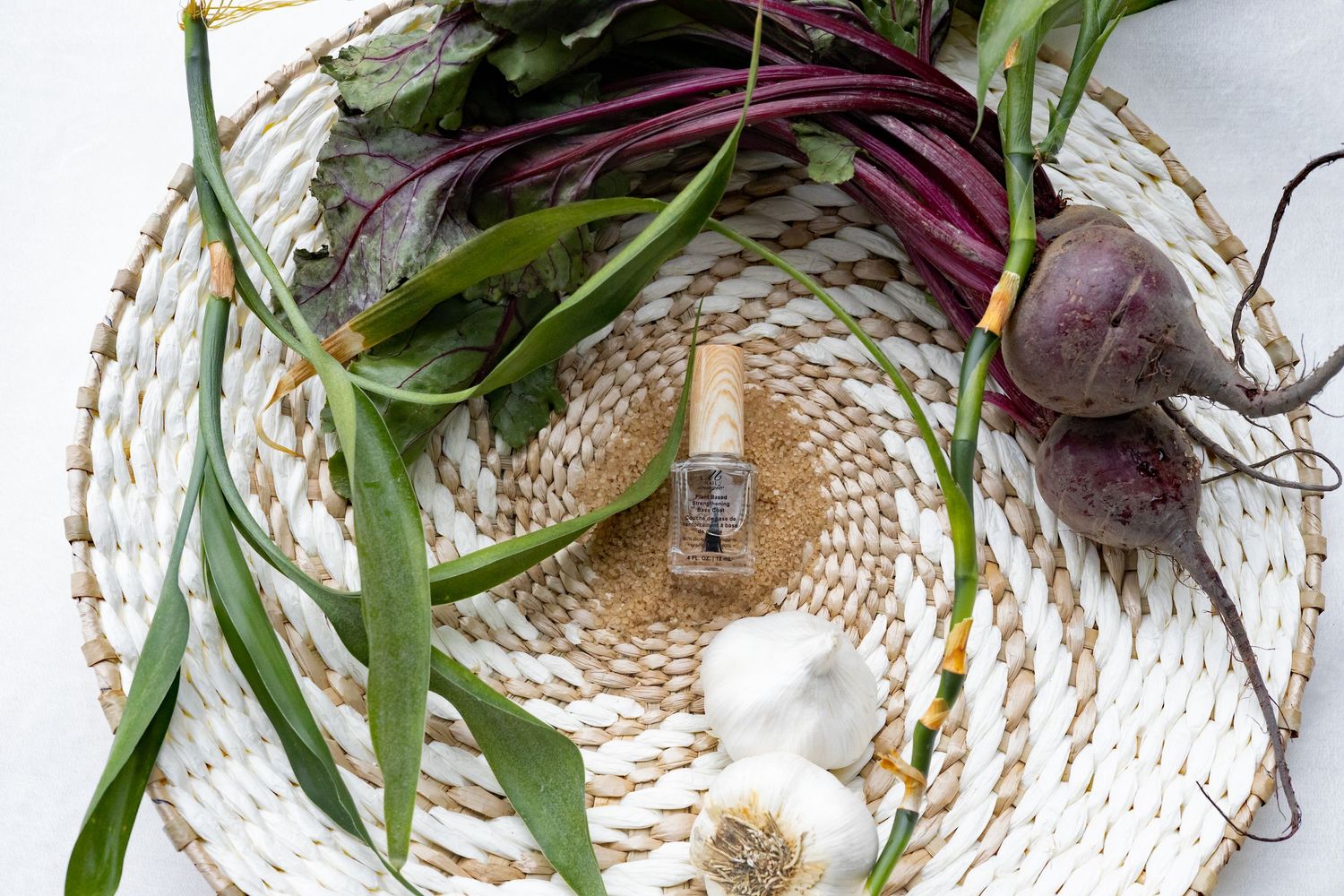 Minimalist styling of nail polish bottle with fresh produce and garlic on woven mat.