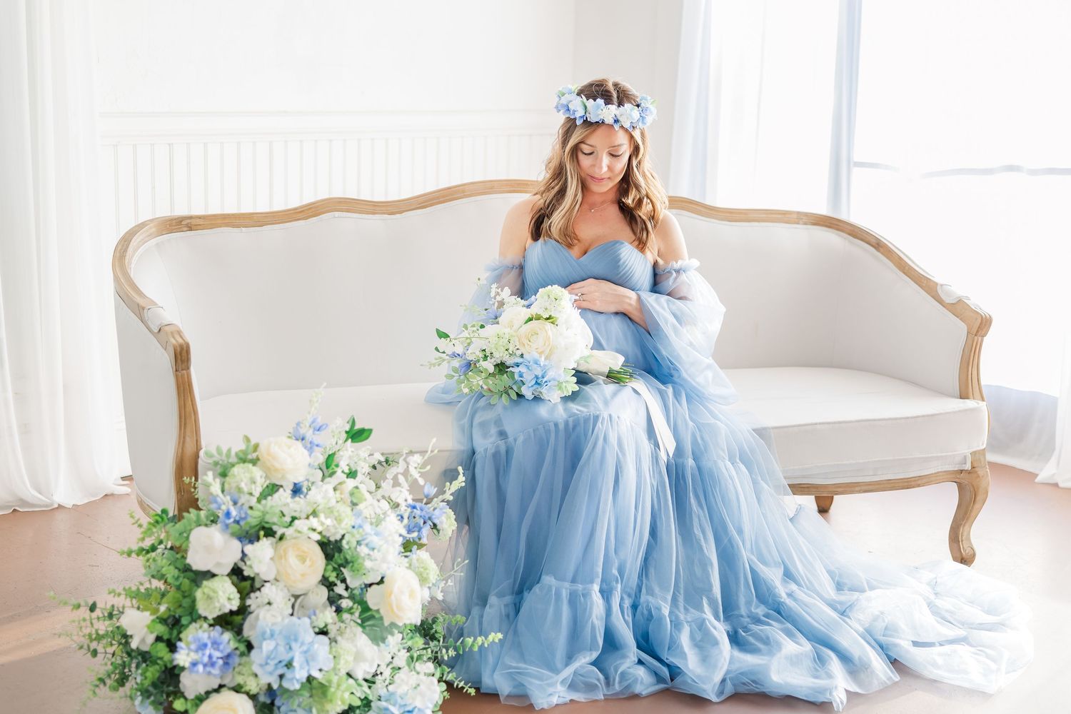 Elegant maternity photo shoot with flowing light blue gown and floral arrangements in a bright white room.