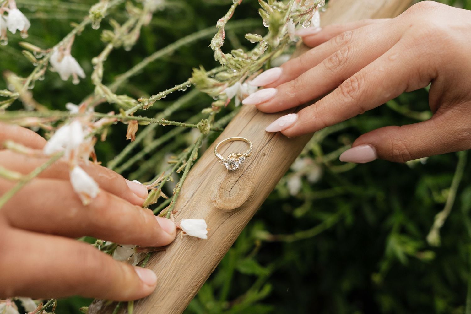 Close-up of hands with white French tip nails touching a diamond engagement ring resting on wood among wildflowers.