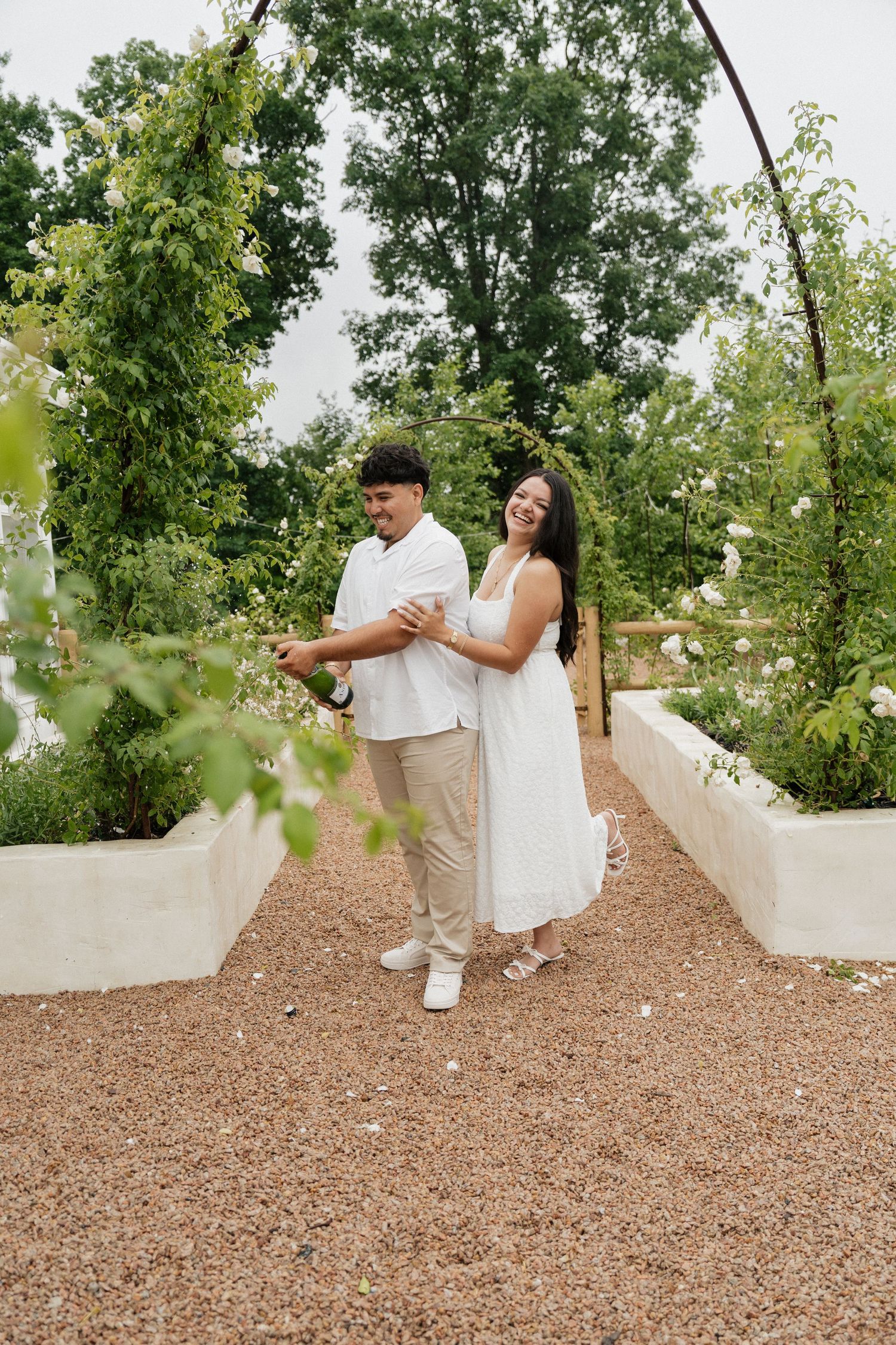 A joyful couple in white attire celebrates under a floral arch in a lush garden, sharing a champagne moment.