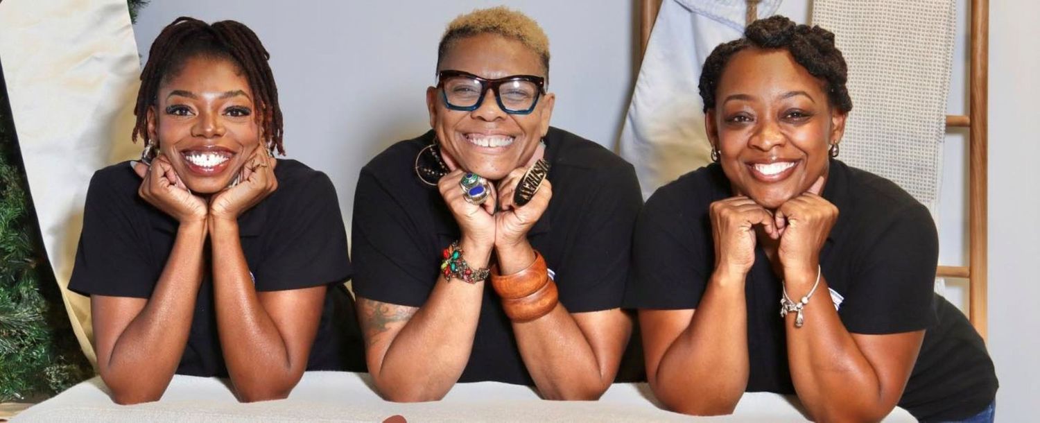Three smiling women in black tops lean on a surface together in a cheerful group portrait photo.