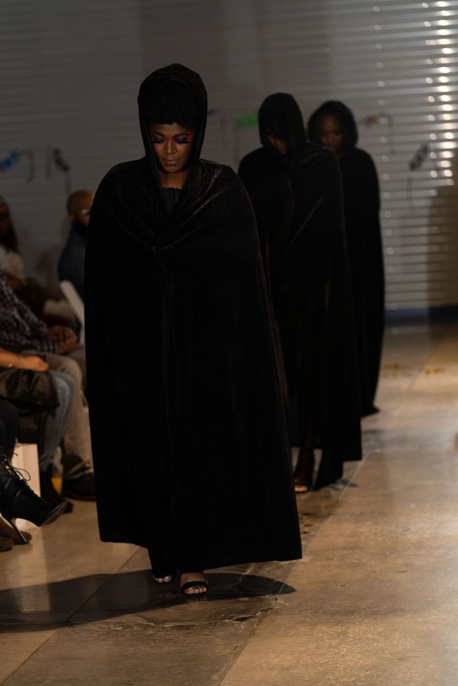 Models in dramatic black velvet hooded cloaks walk runway at fashion show.
