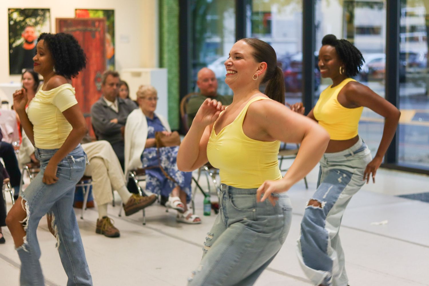 Three dancers in matching yellow tops and jeans perform lively routine at indoor event.