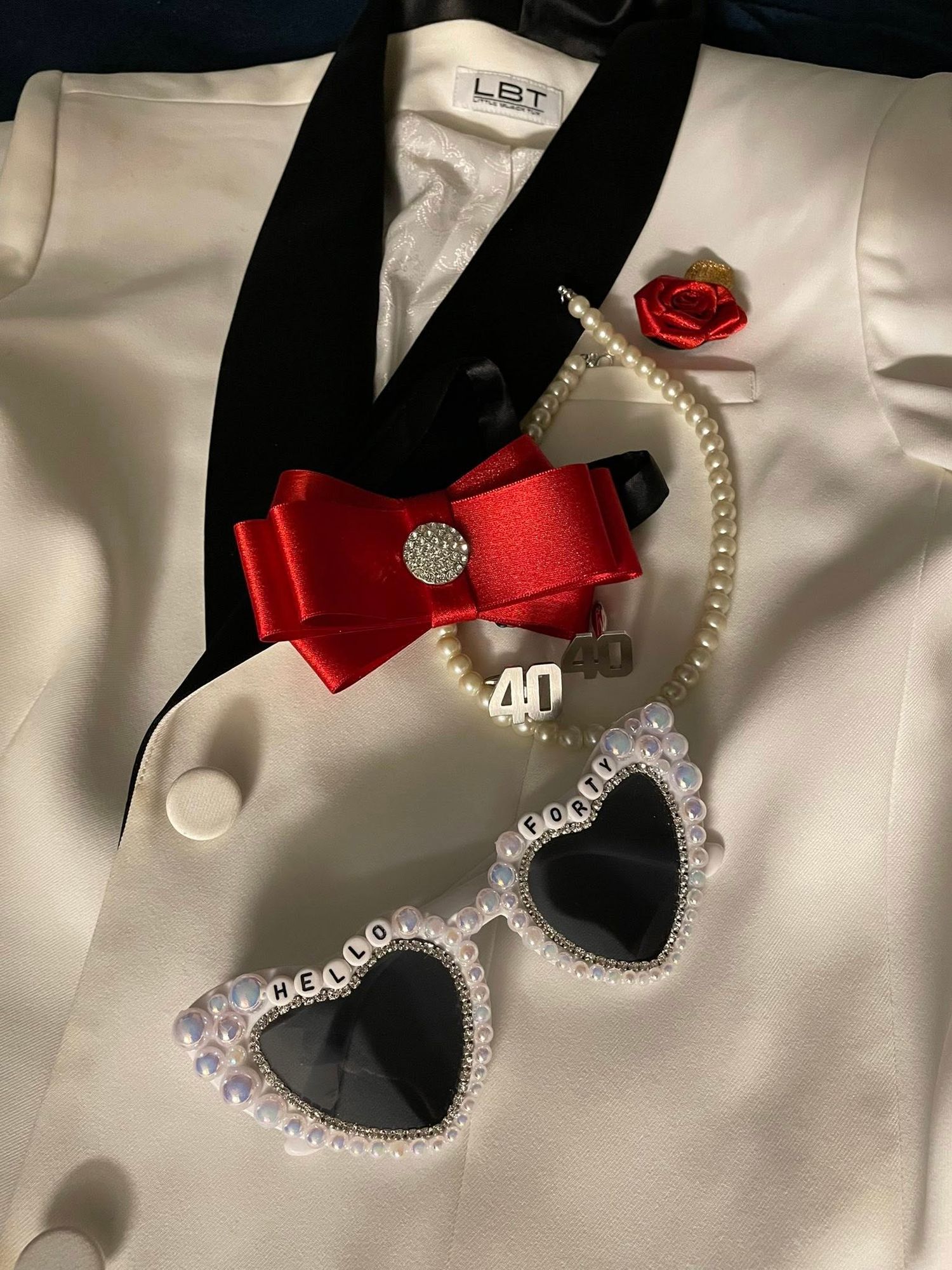 A white blazer with red bow, pearl bracelet, 40 charm, and heart-shaped pearl sunglasses.