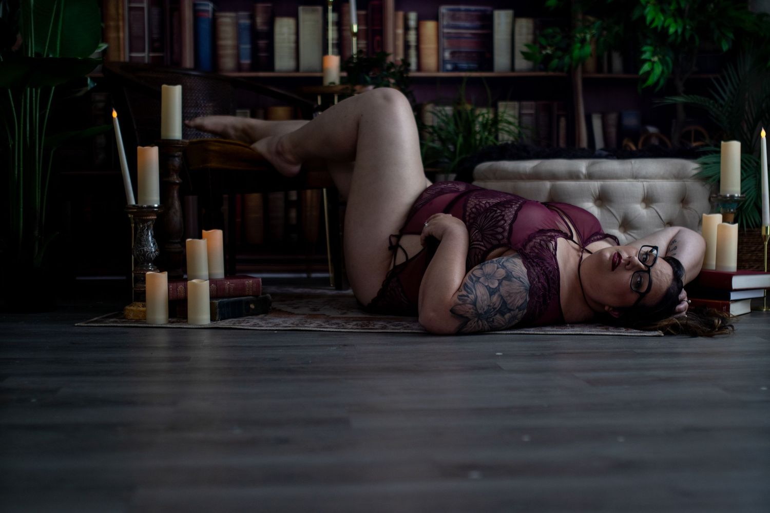 Woman in purple lingerie posing on floor surrounded by candles and books in moody setting.