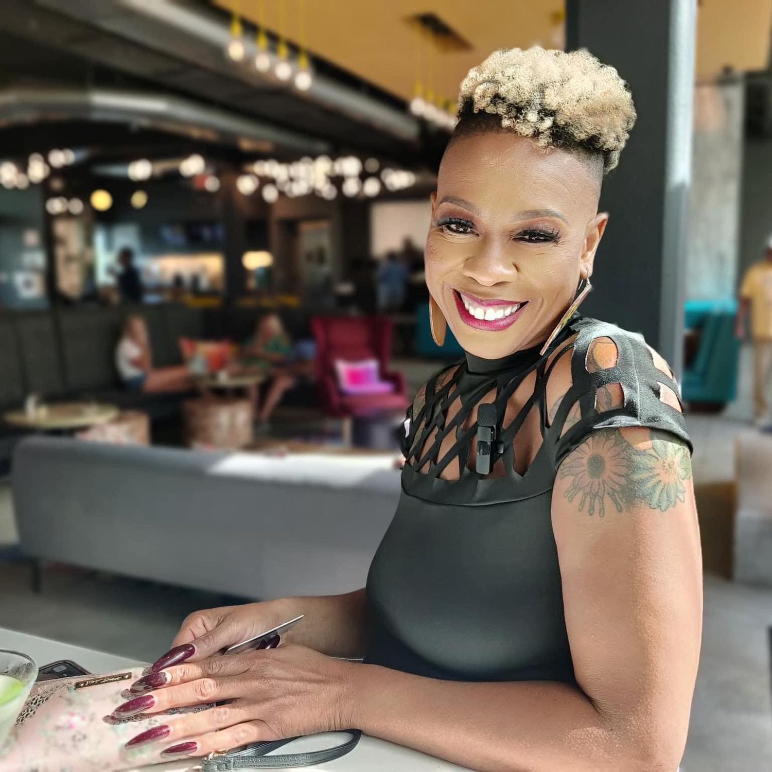 A smiling woman with blonde natural hair and tattoos sits at a table in a modern restaurant.