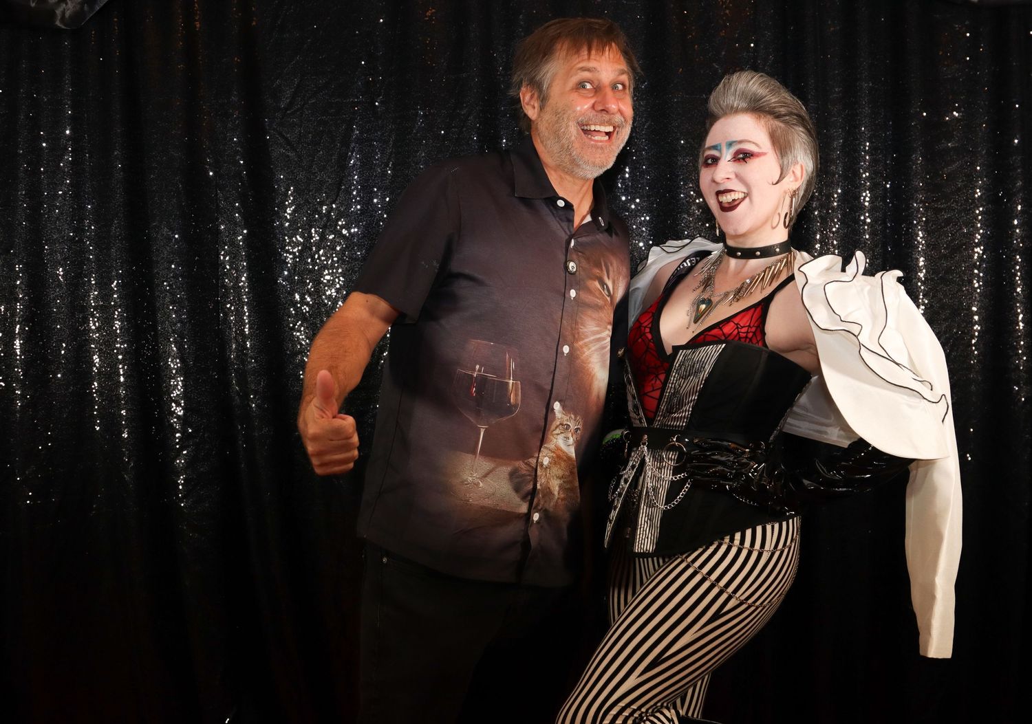 Man and woman in costume laugh together in front of a black sequin backdrop.