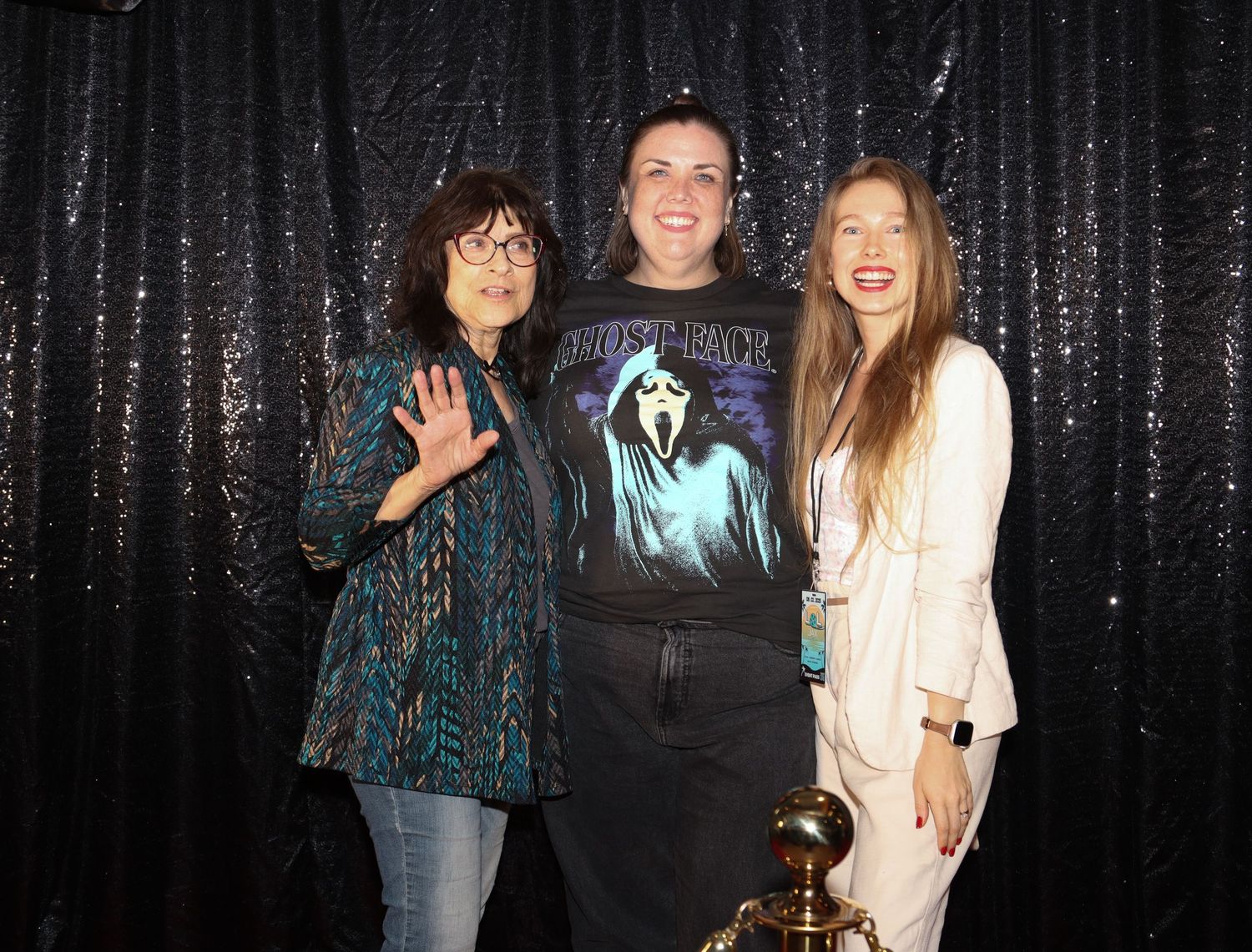 Three women pose together in front of a black sequin backdrop at an indoor event.