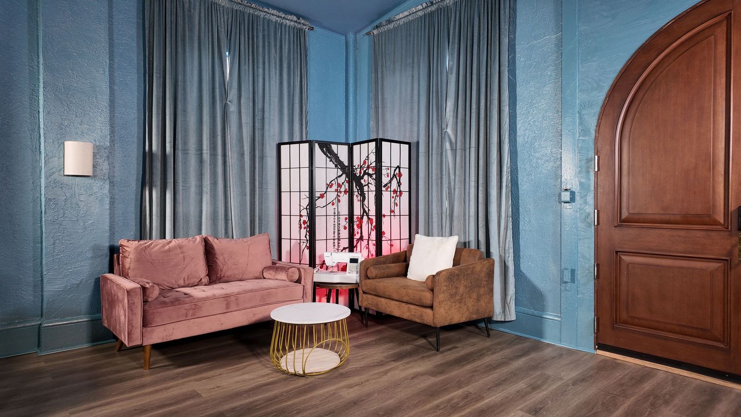 An elegant lounge room with blue textured walls, pink velvet sofa, brown armchair, and an arched wooden door.