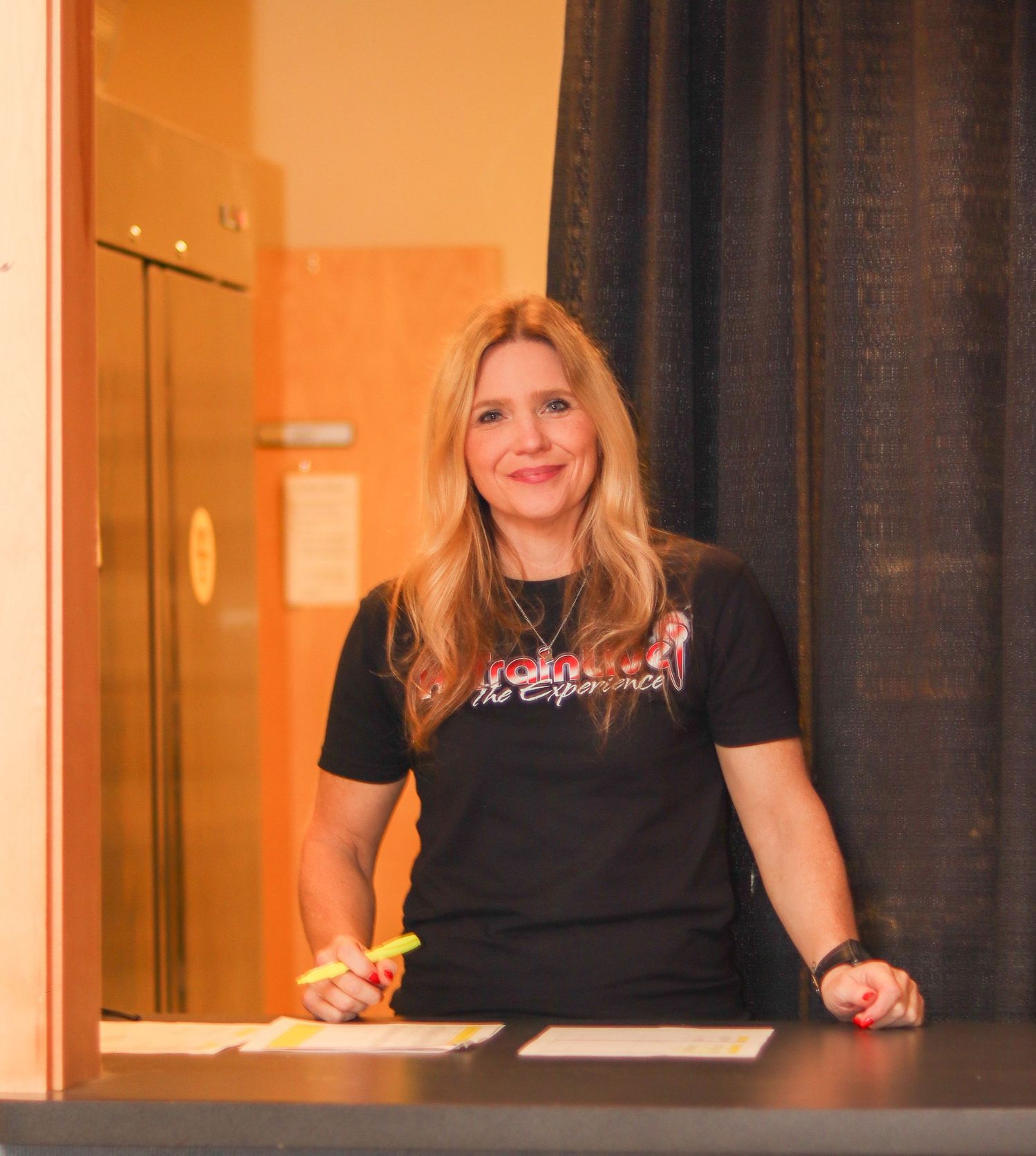 Smiling blonde woman in black t-shirt working at a registration or check-in desk.