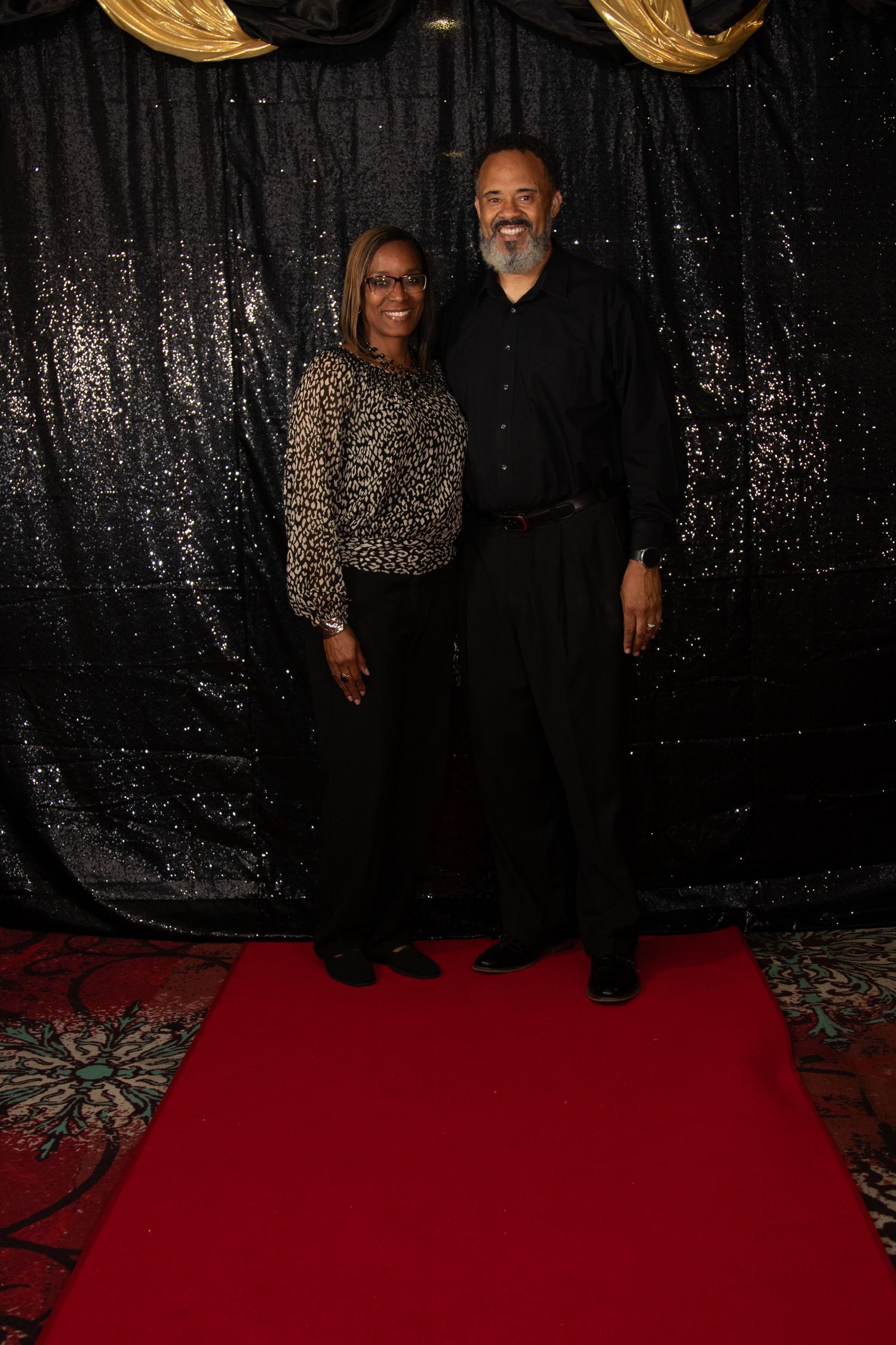 A smiling couple poses together on a red carpet in front of a black sequin backdrop.