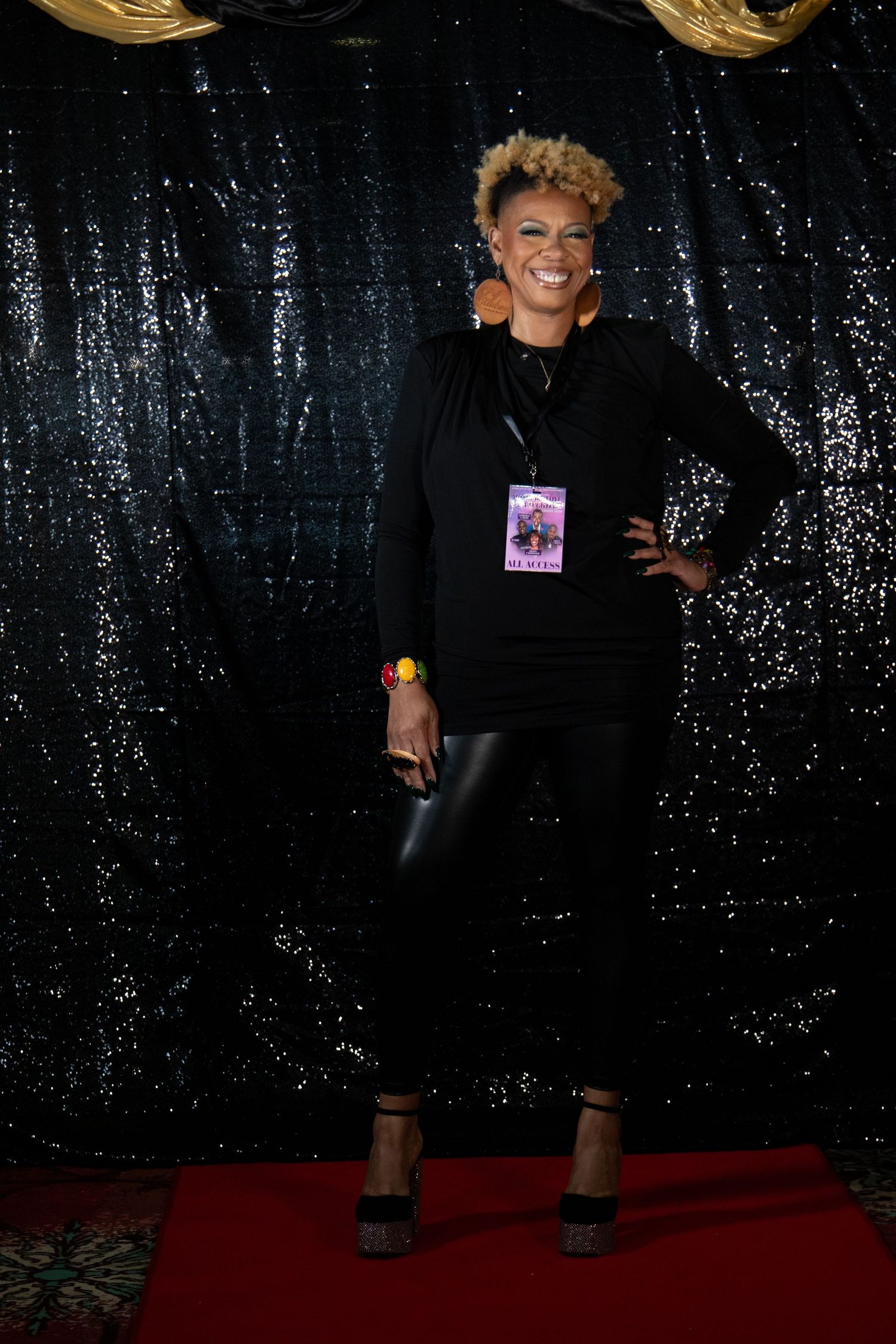A woman in all-black outfit with blonde curly hair poses on a red carpet with sequin backdrop.