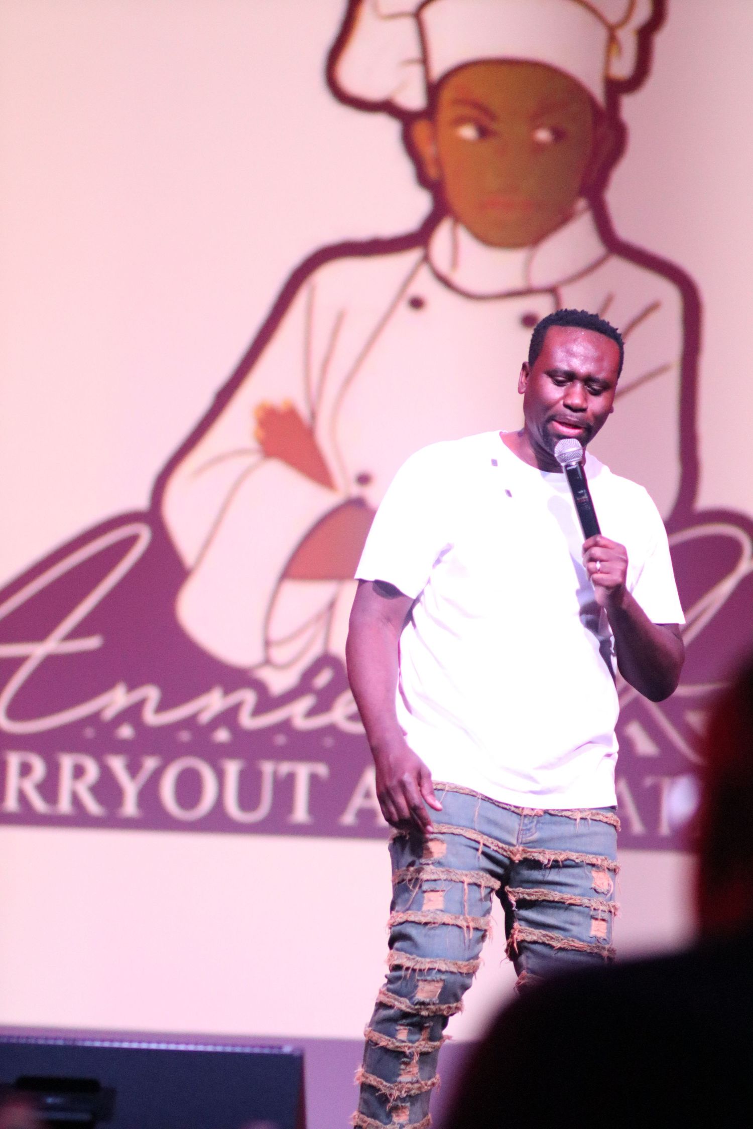 Male performer in white tee and ripped jeans speaking into mic at Annie's Carryout event.