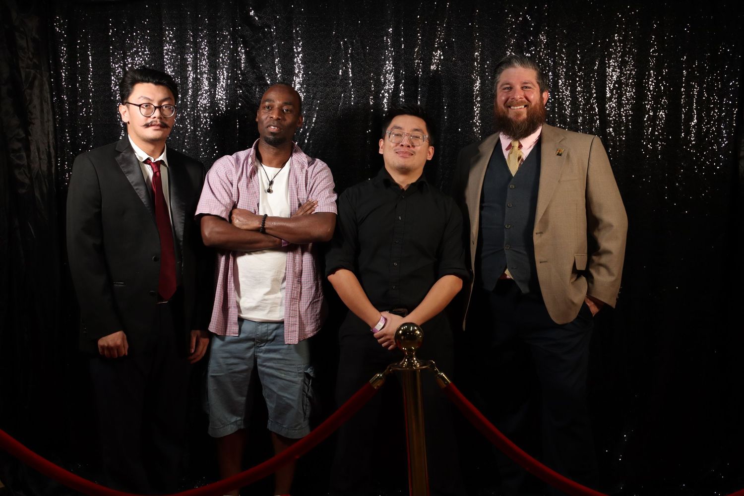 Four men posing together in front of a black sequin backdrop at a formal event.