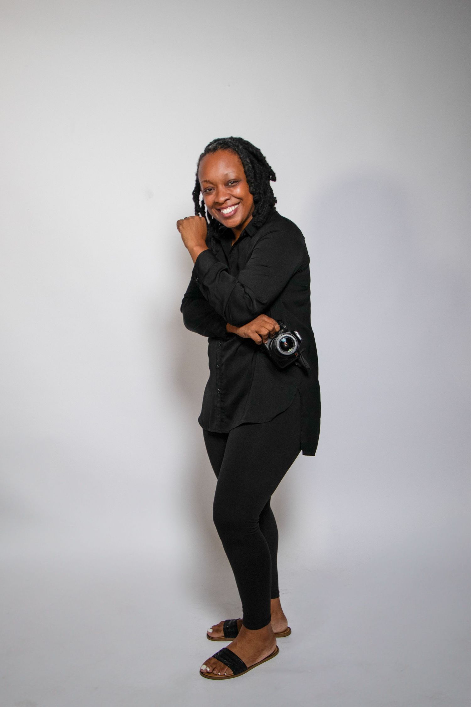 A smiling woman in all-black outfit holds a camera while posing against a white background.