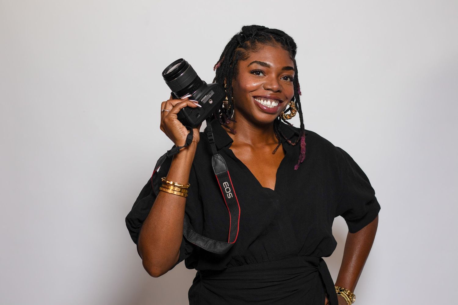 A joyful woman in black holds a DSLR camera on her shoulder, wearing gold jewelry.
