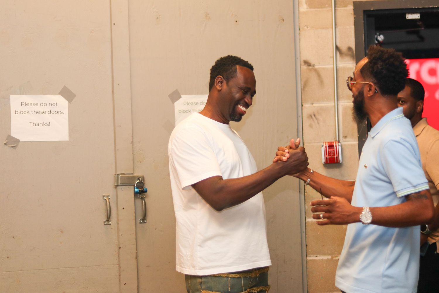 Two Black men share a friendly handshake and laugh together near a metal door indoors.
