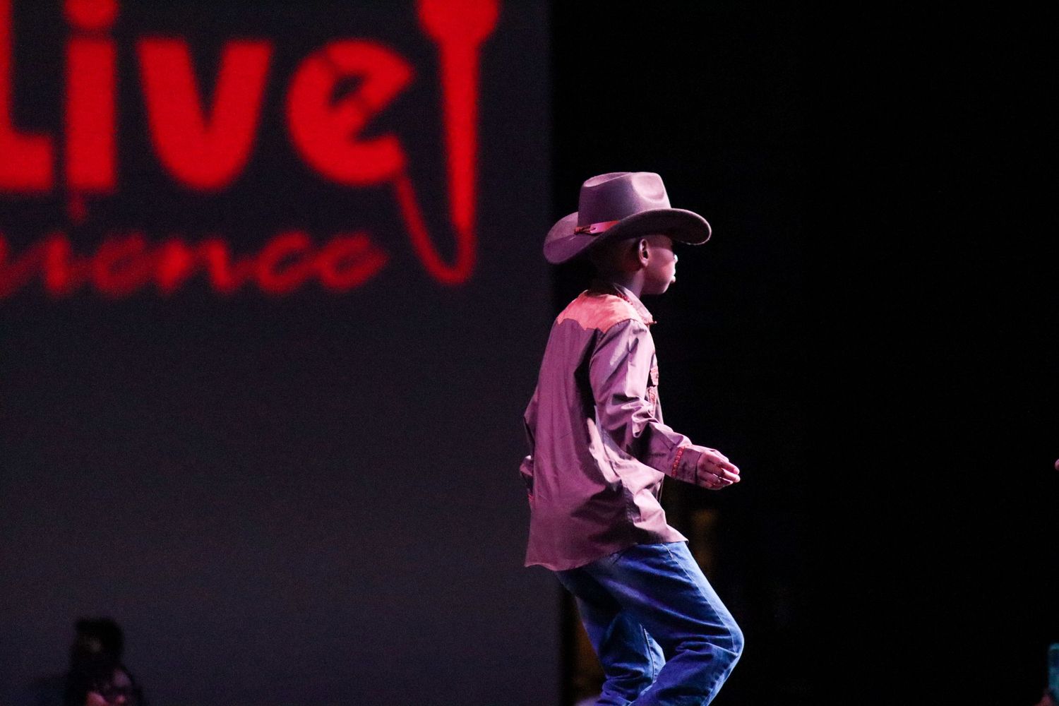 A performer in a pink cowboy hat and shirt walks across a stage at A-Train Live.