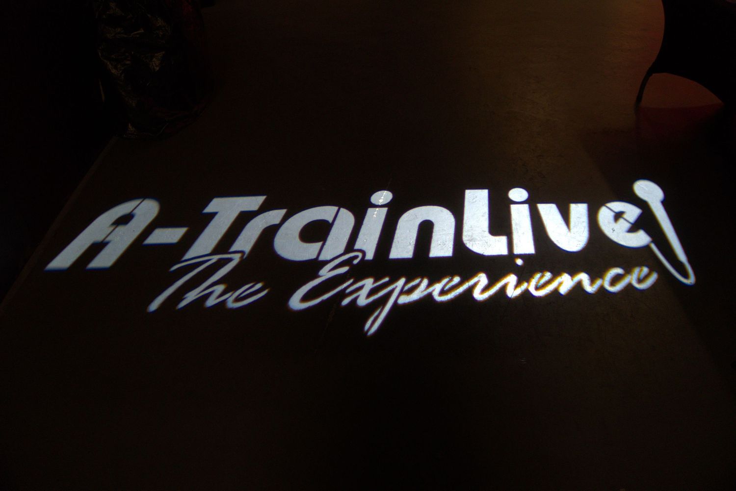 A-Train Live The Experience logo projected in silver light on a dark surface.