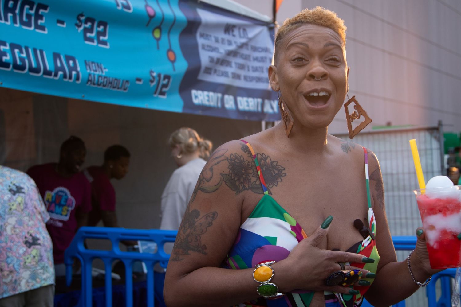 Tattooed woman smiling and holding snow cone at an outdoor fair or festival.