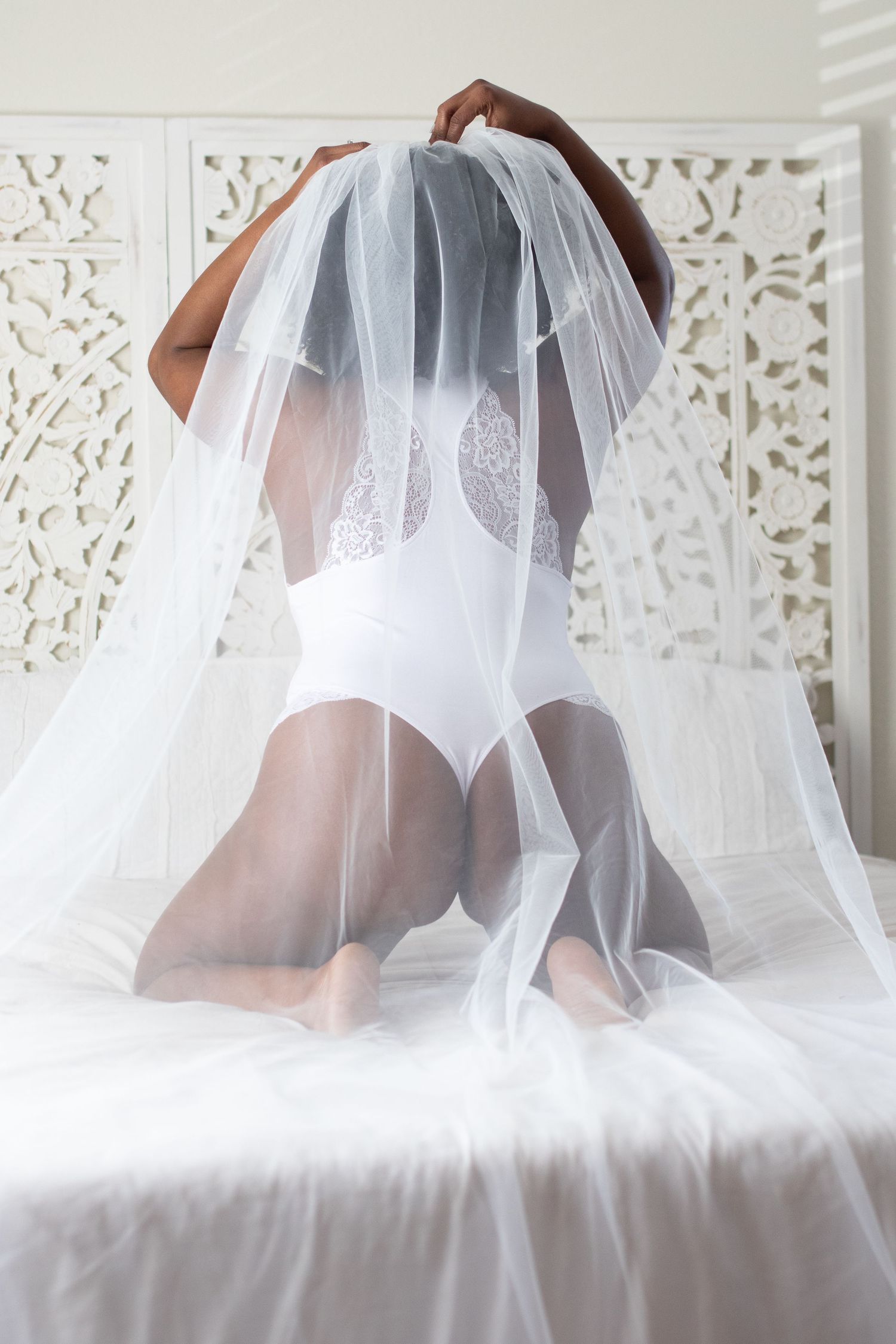 Person in white lace bodysuit kneeling under a flowing veil against decorative white screen.