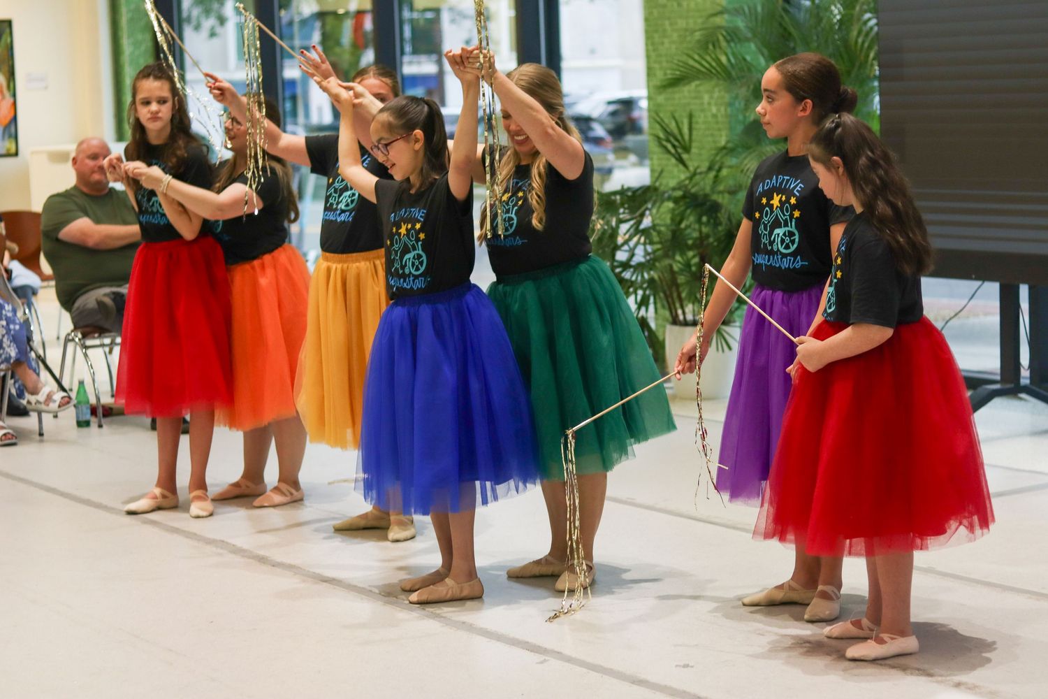Young performers in colorful tulle skirts and black tops rehearse dance routine with props.