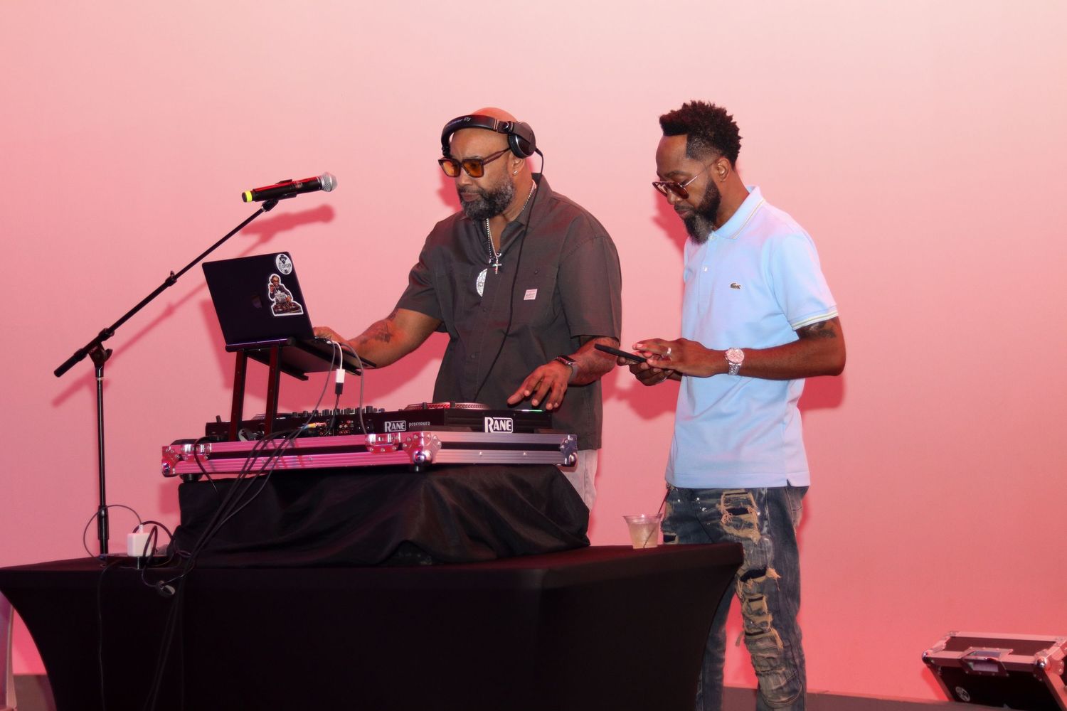 Two men working at a DJ booth with turntables against a pink backdrop at an event.