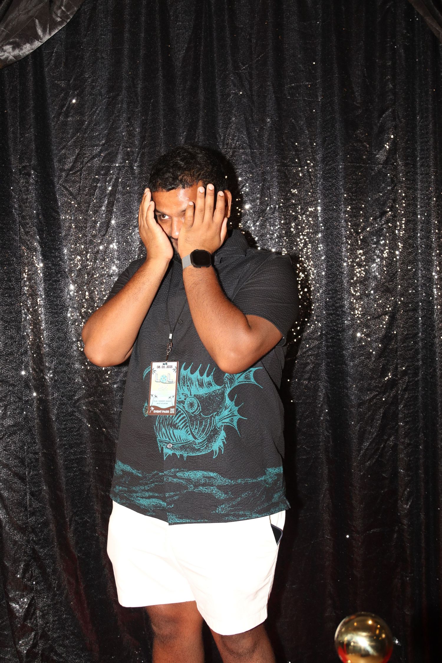Young man covering his face with hands standing in front of a black sequin backdrop.