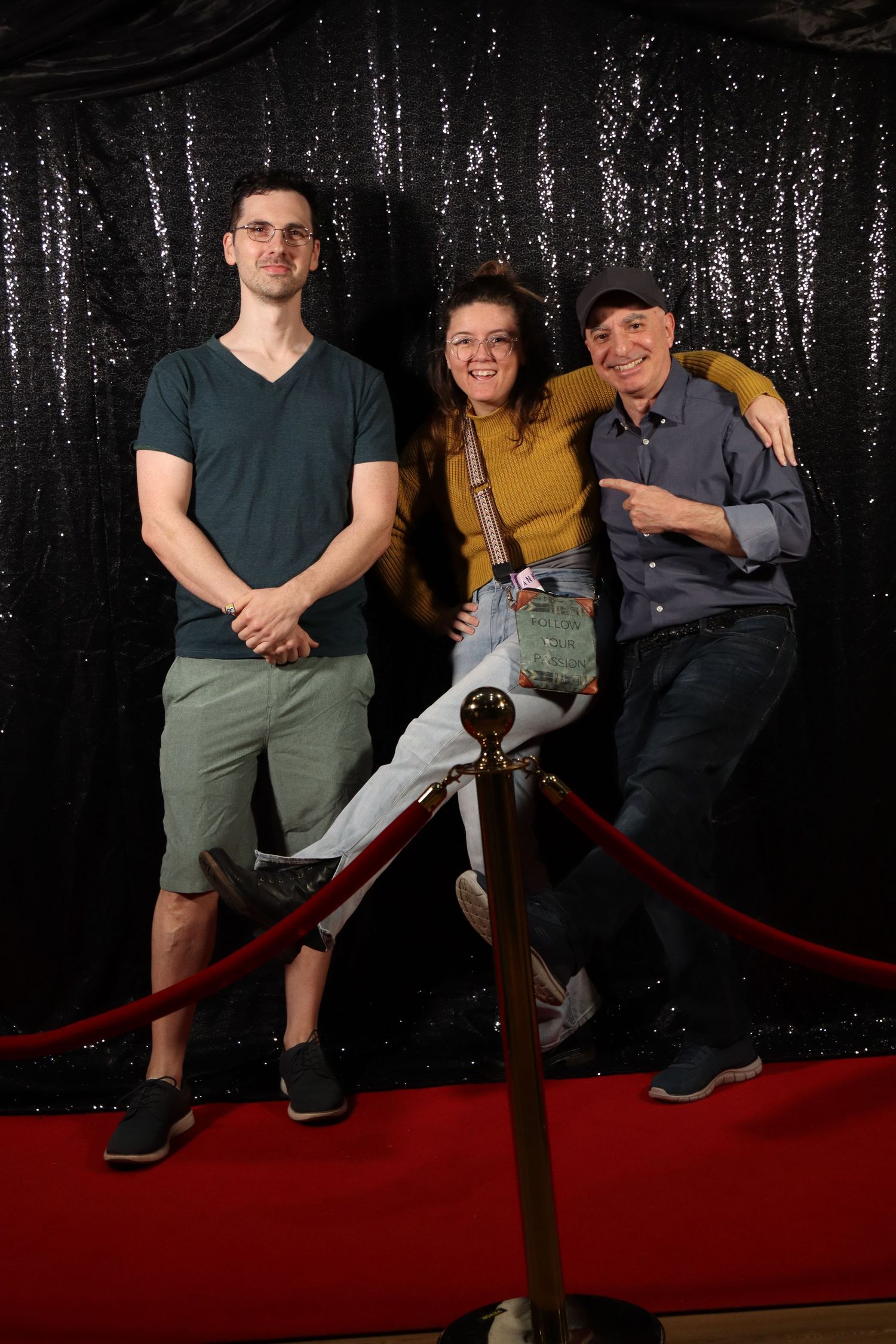 Three people smile together on a red carpet in front of a black sequin photo backdrop.