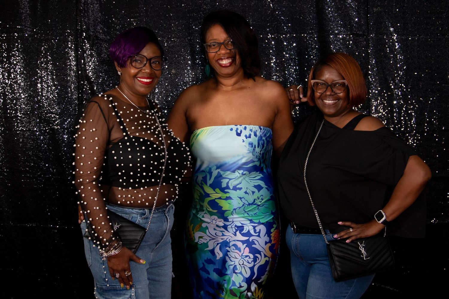 Three women smiling together in front of a sparkling black sequin backdrop at an event.