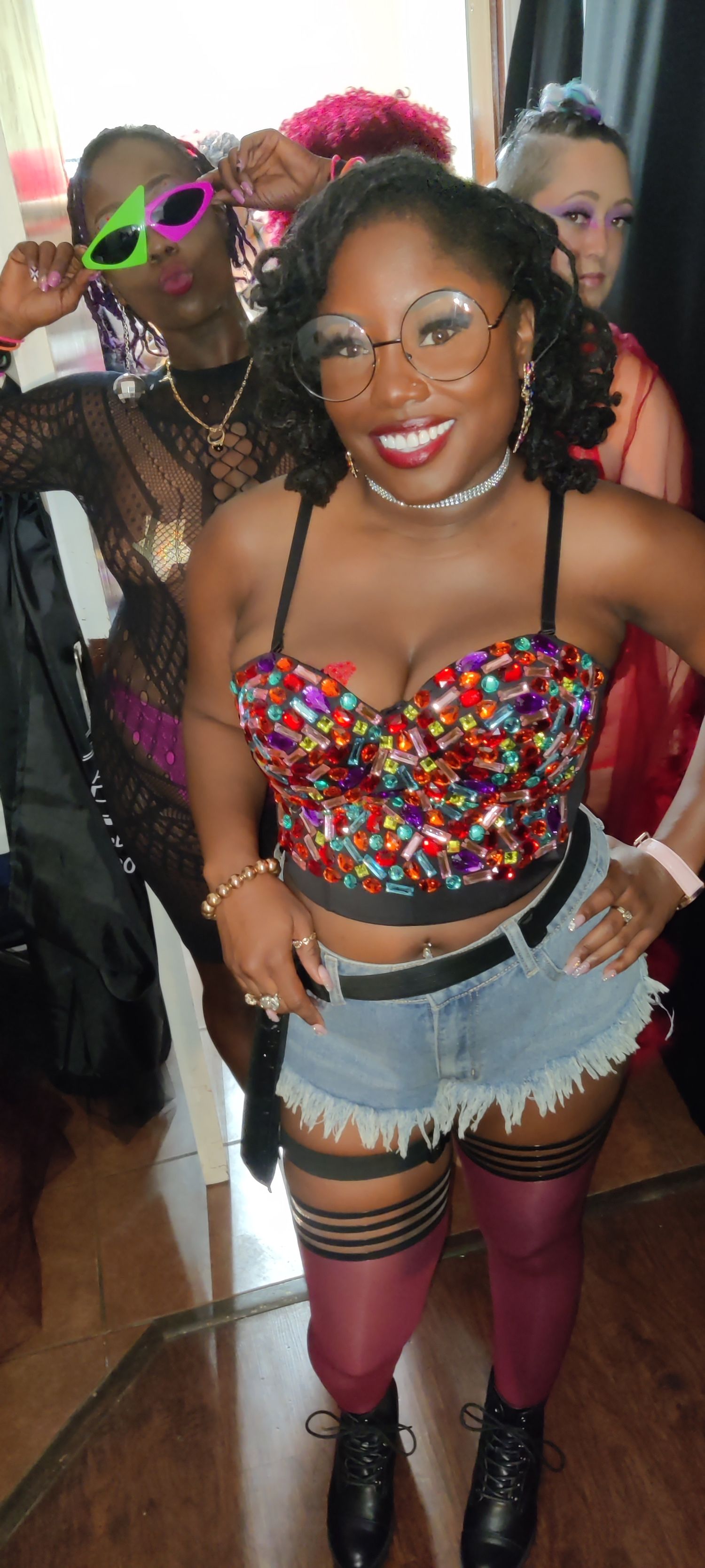 A smiling Black woman wearing a colorful jeweled crop top, denim shorts, thigh-high stockings, and round glasses at a party.