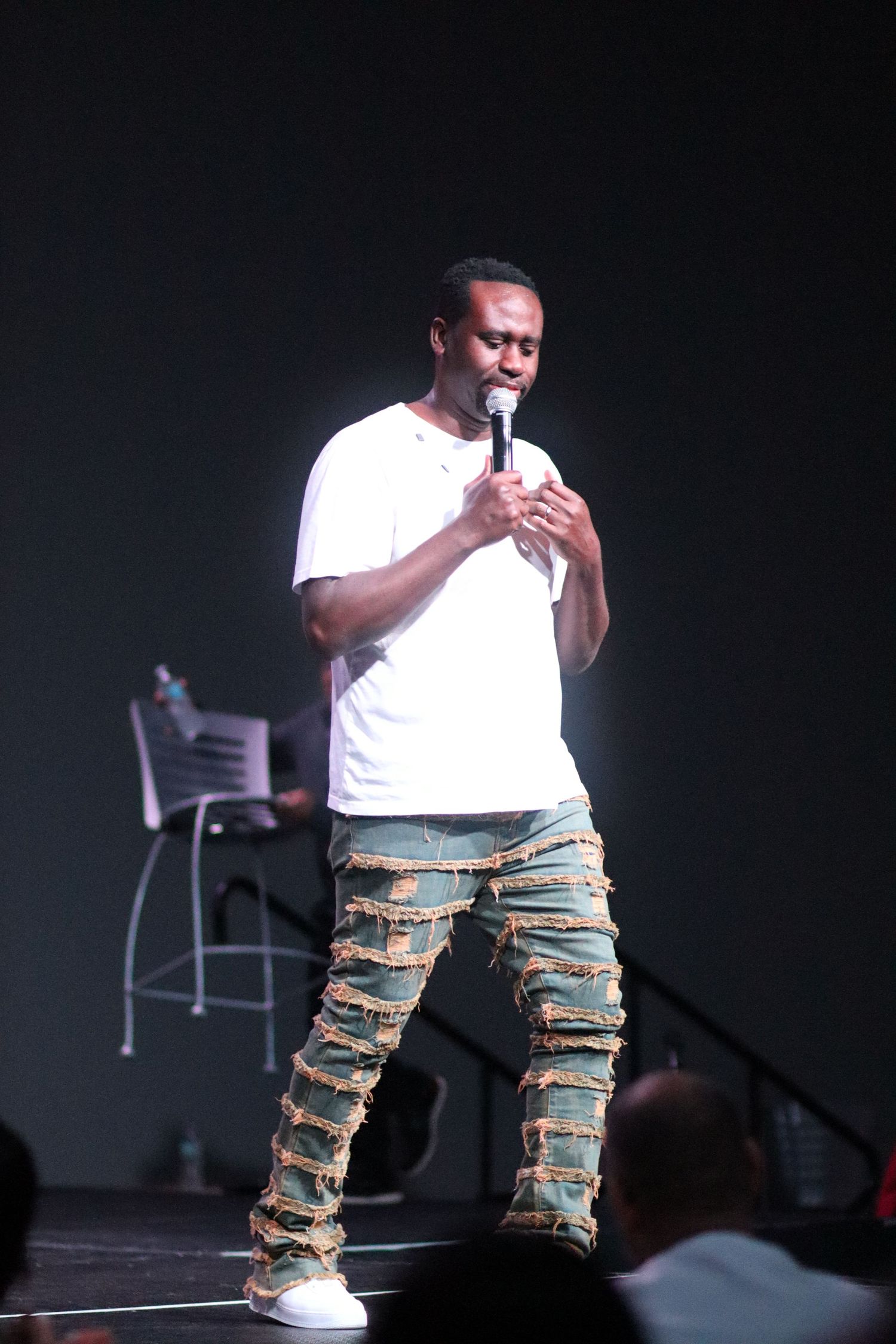 A man in a white t-shirt and distressed jeans performs stand-up comedy on a dark stage.