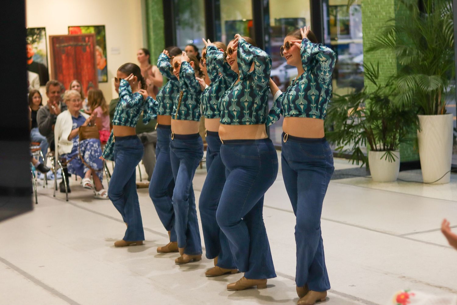 Line of female dancers in jeans and plaid shirts performing a country line dance routine on an indoor stage.