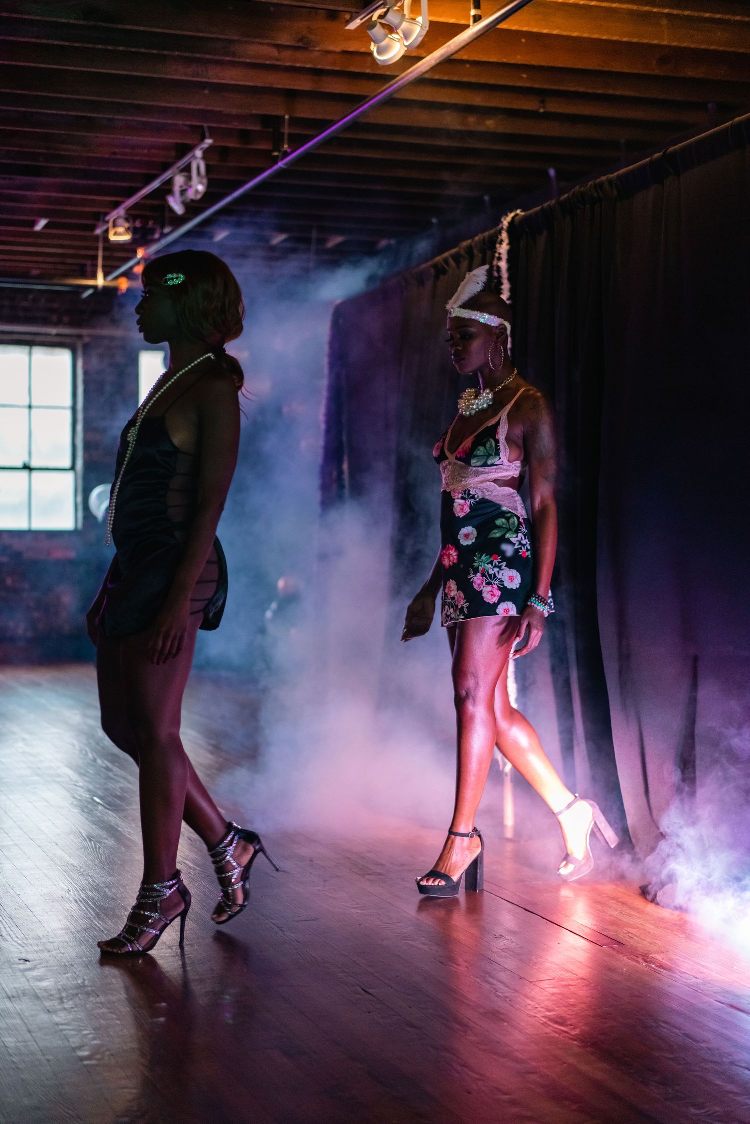 Two female models pose dramatically in smoky purple-lit fashion show venue.