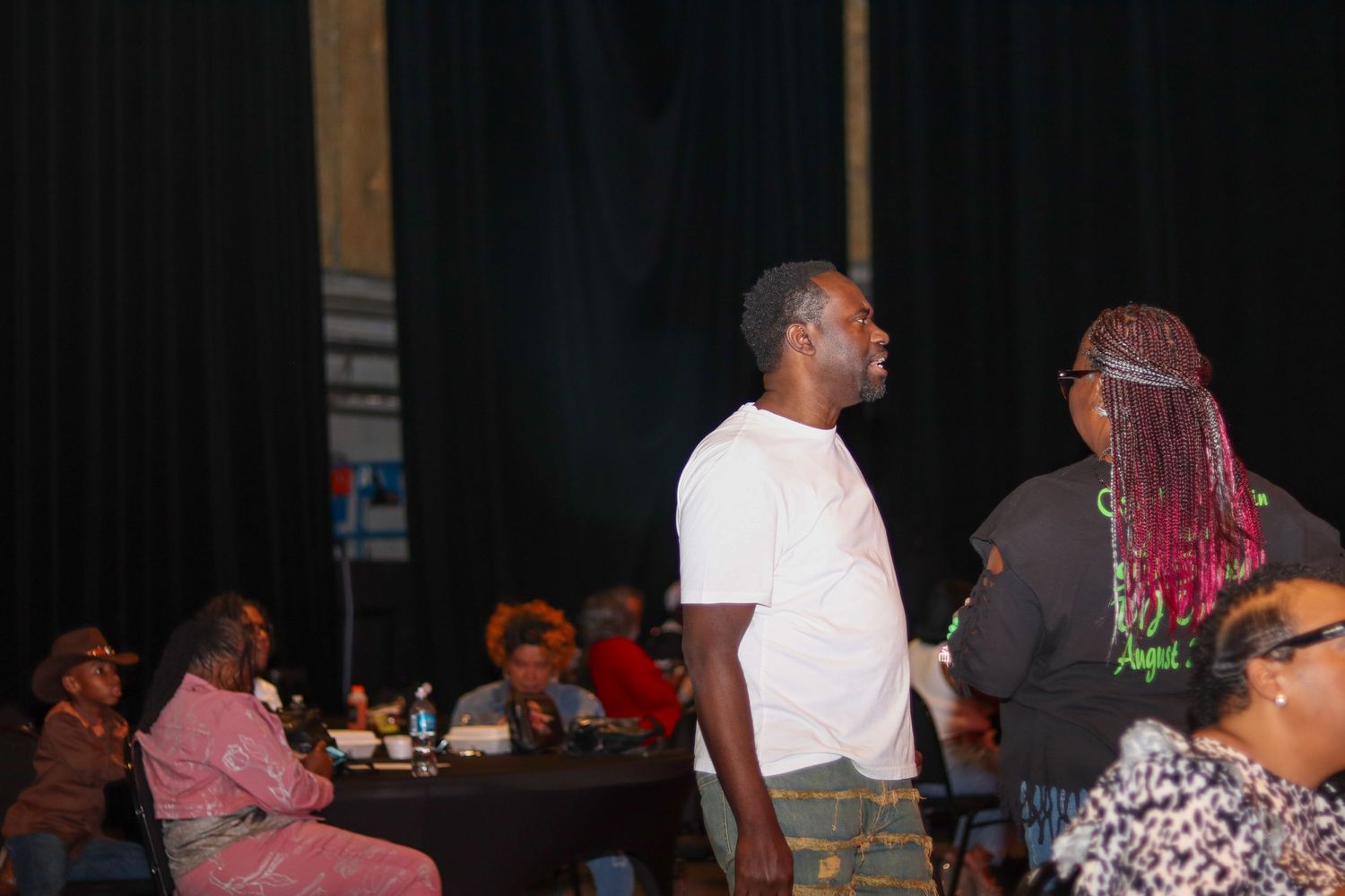 Man in white t-shirt standing at an event venue with seated audience members at tables.