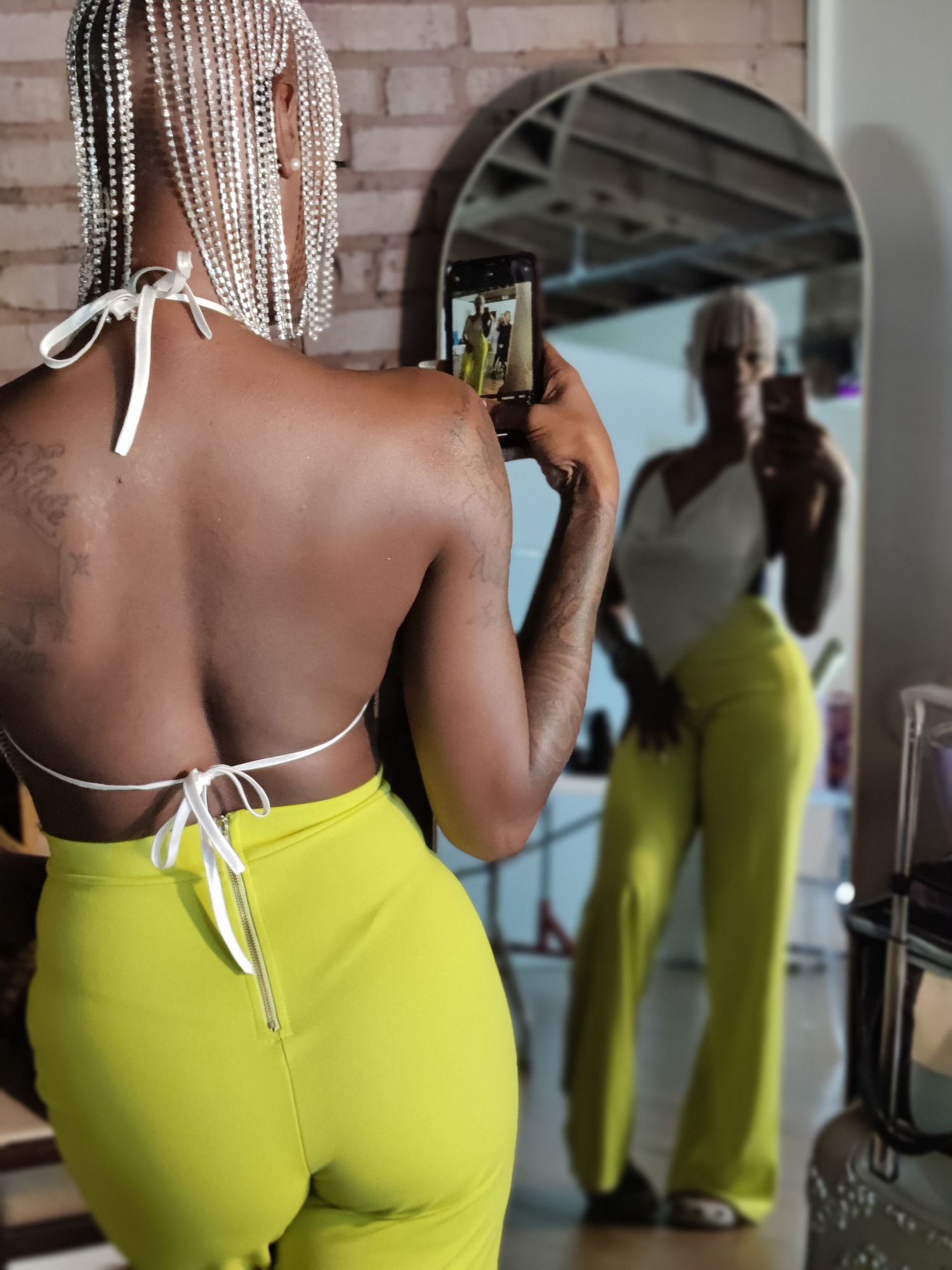 Woman with white braids photographs herself in a mirror wearing a yellow-green backless outfit.