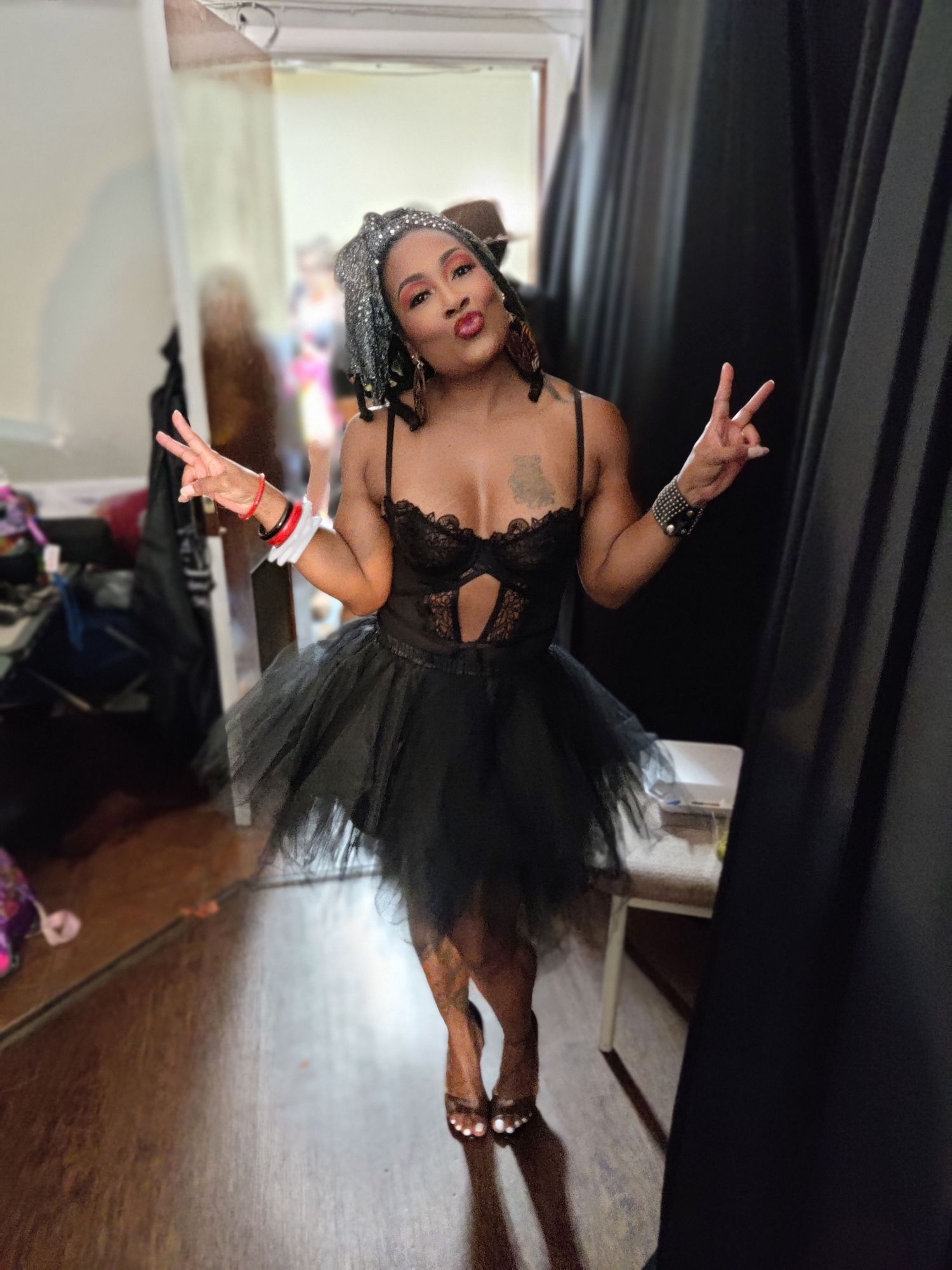 A Black woman in a black lace bodysuit and tutu skirt poses with peace signs in front of a mirror.