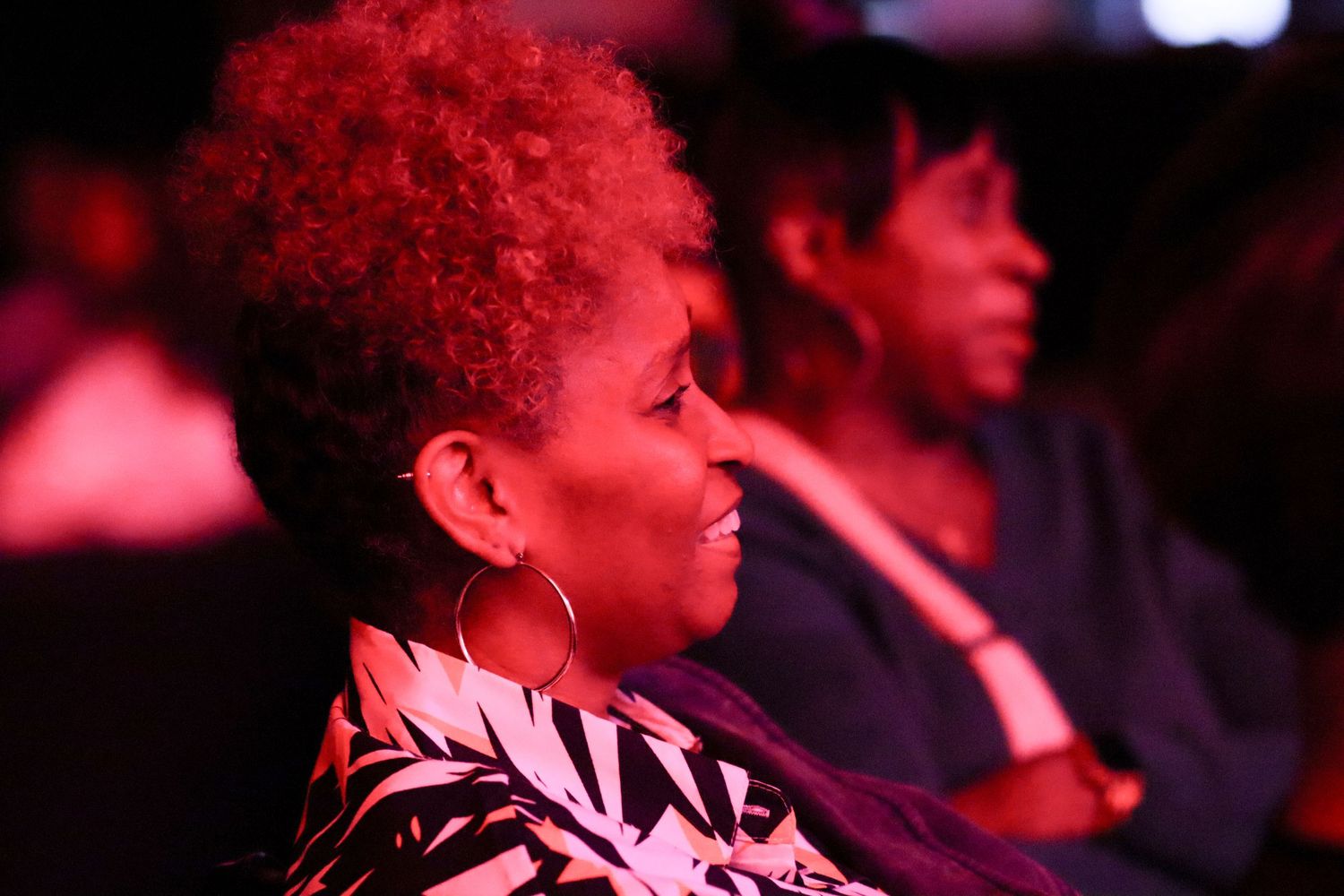A woman with red hair and hoop earrings watches a performance in a dimly lit venue.