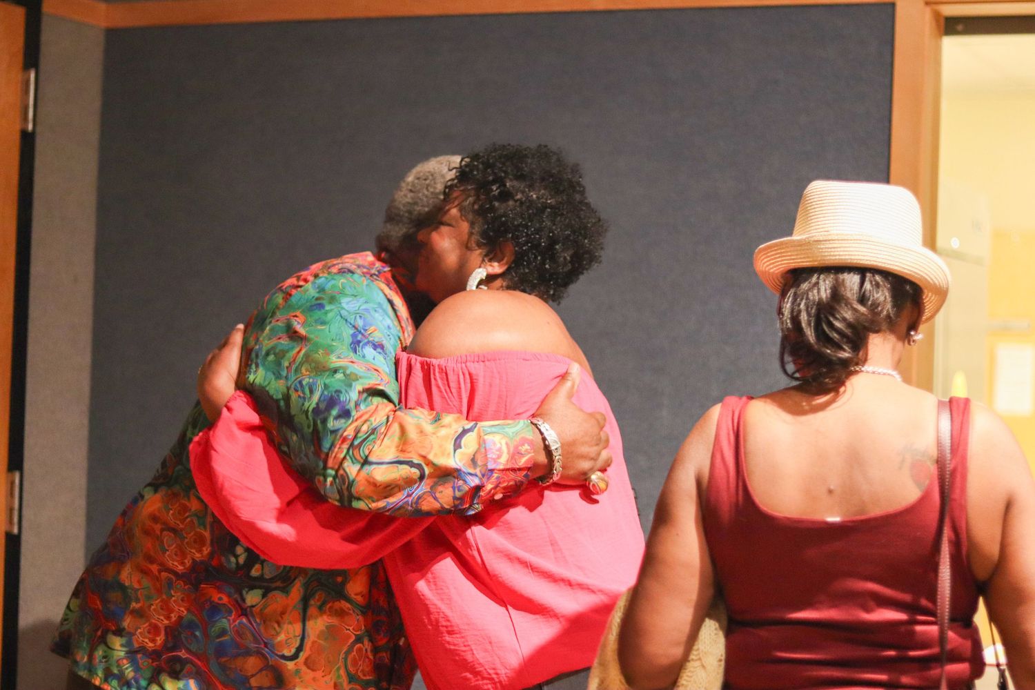 Two women share a warm embrace while a third woman in a white hat stands nearby indoors.