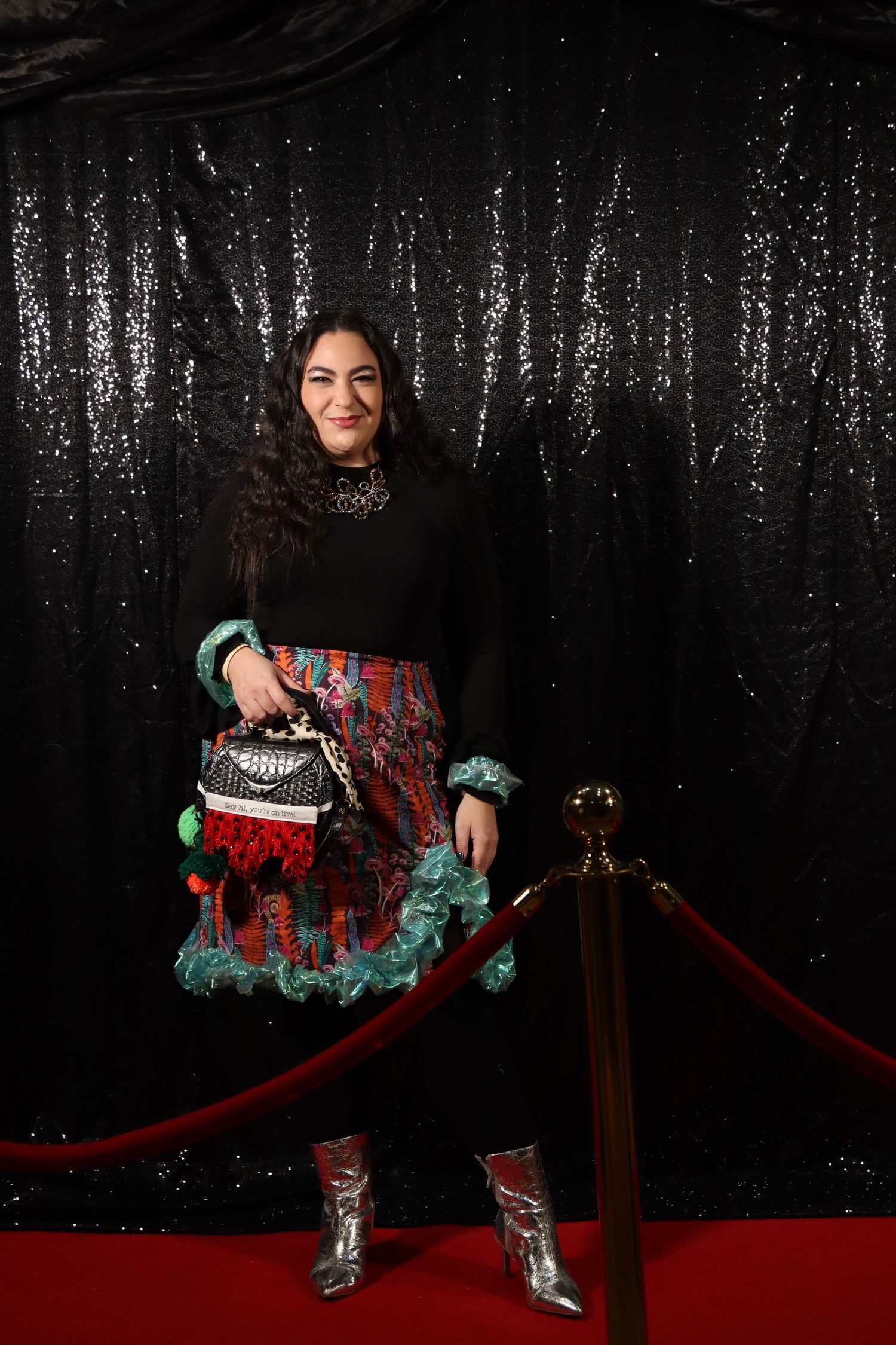 Woman in colorful skirt and silver boots poses on red carpet with black sequin backdrop.