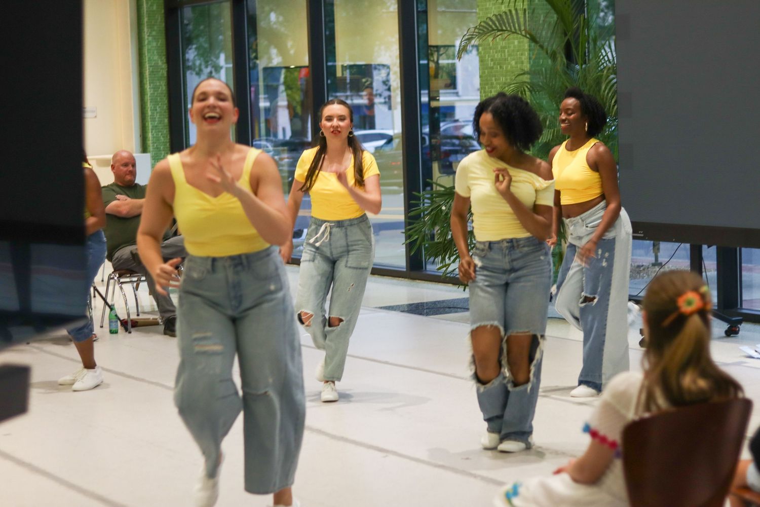 Three women in yellow tops and jeans perform energetic dance routine at Bridges event.