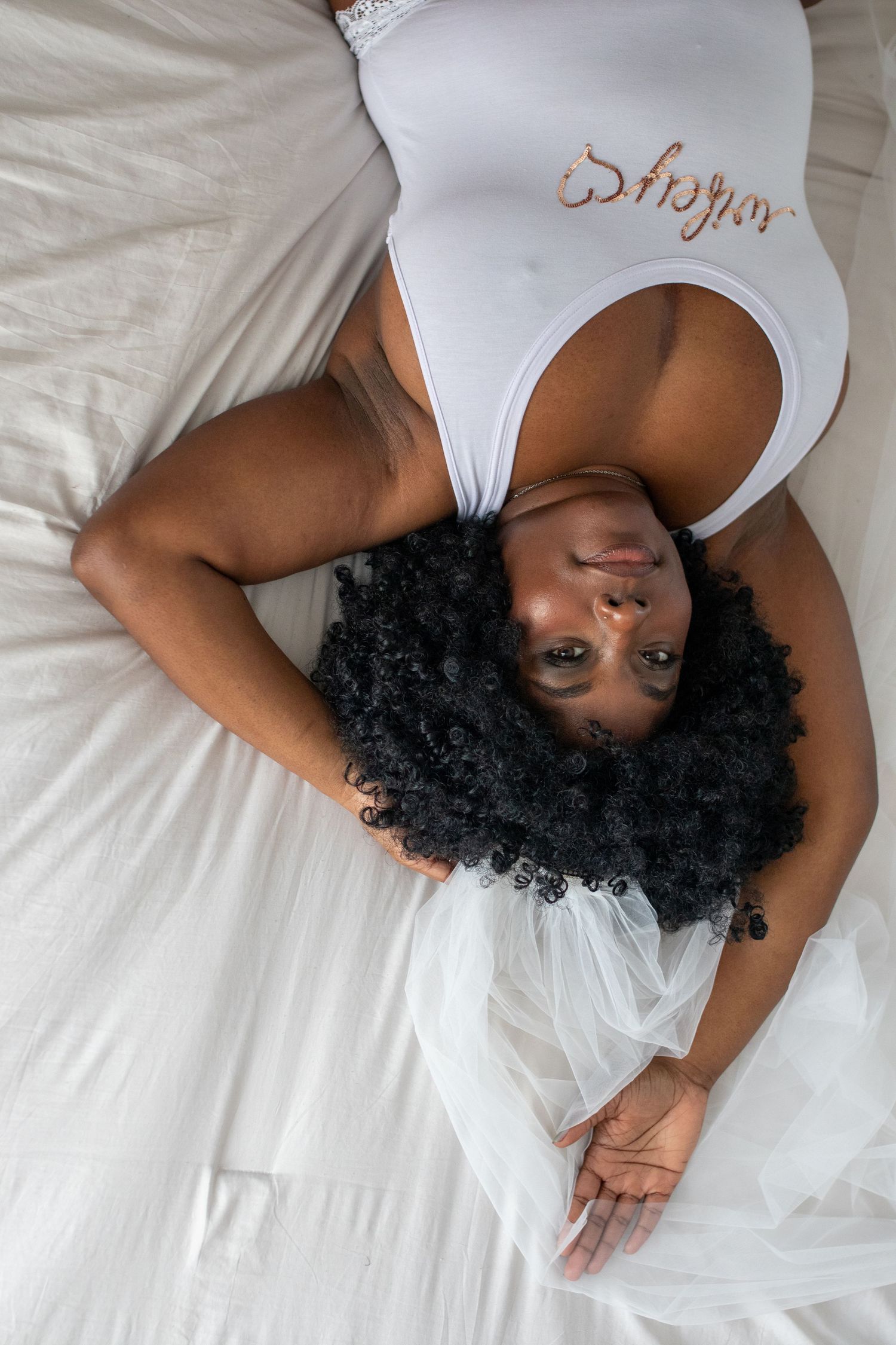 Woman in white bodysuit lying on bed photographed from above with curly black hair spread out.
