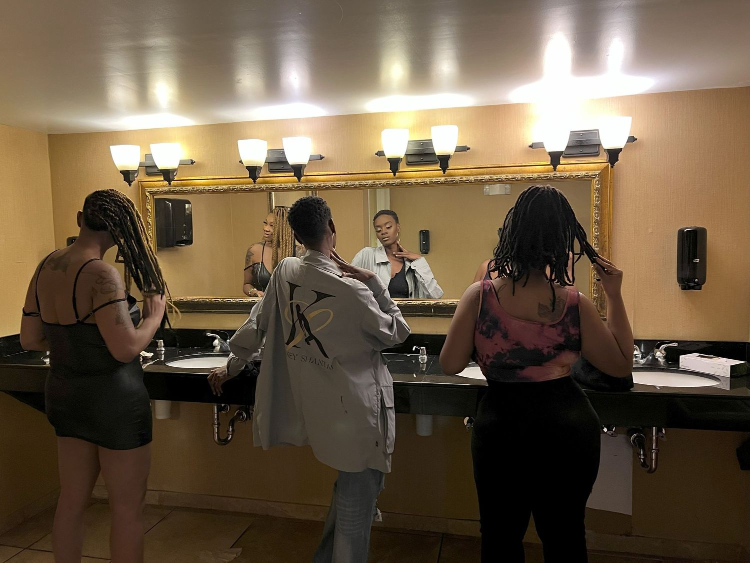 Three women with locs style their hair in front of a large mirror in a well-lit bathroom.
