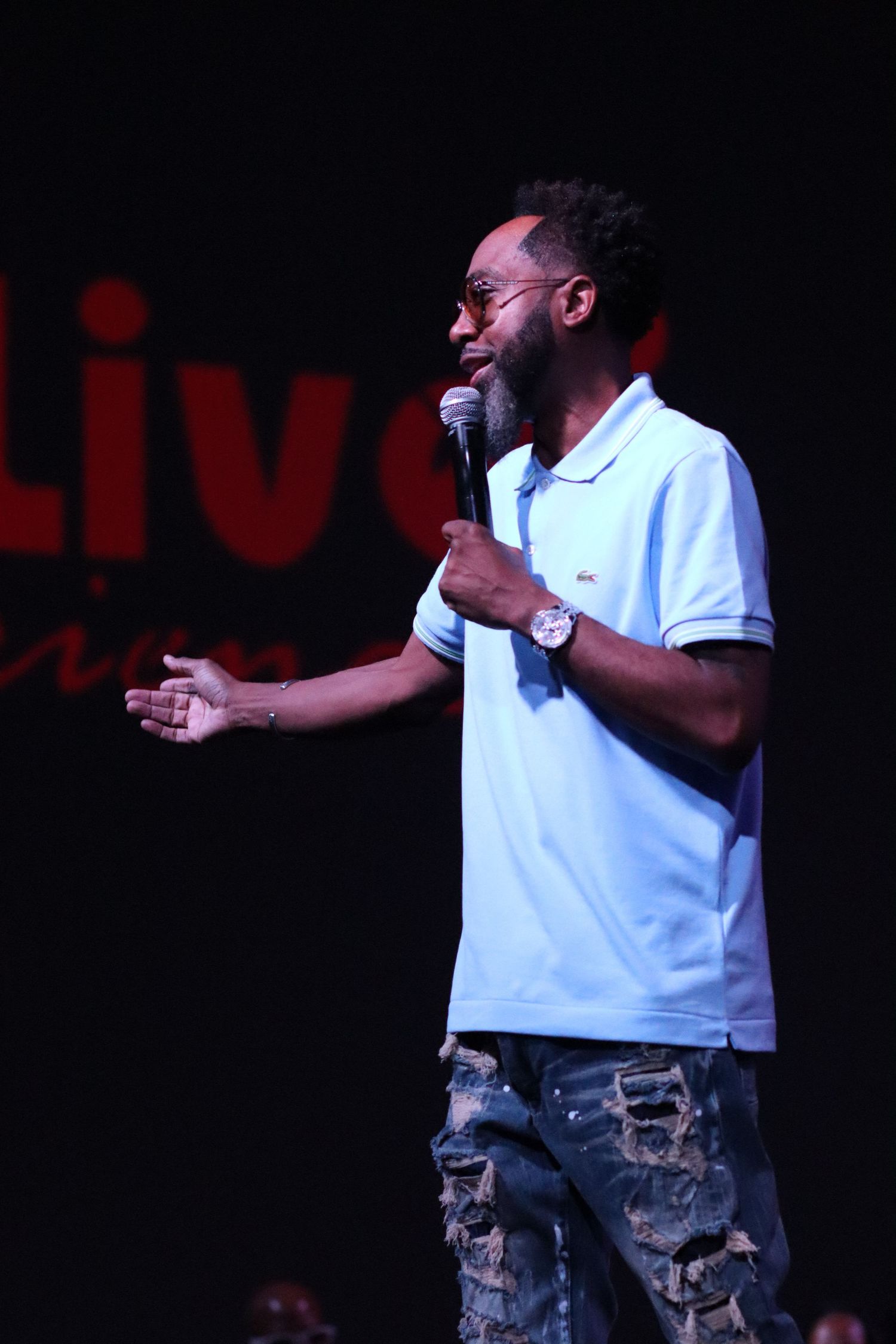 Stand-up comedian in white polo shirt performs with microphone on dark stage.