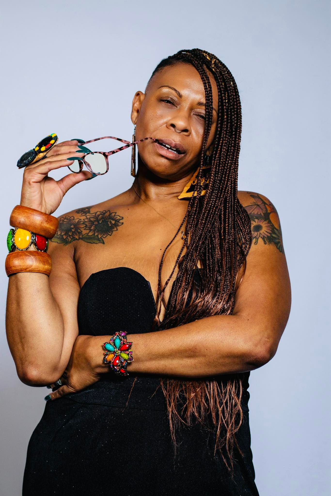 A confident woman with long braids, colorful bracelets, and tattoos poses in a black strapless top.